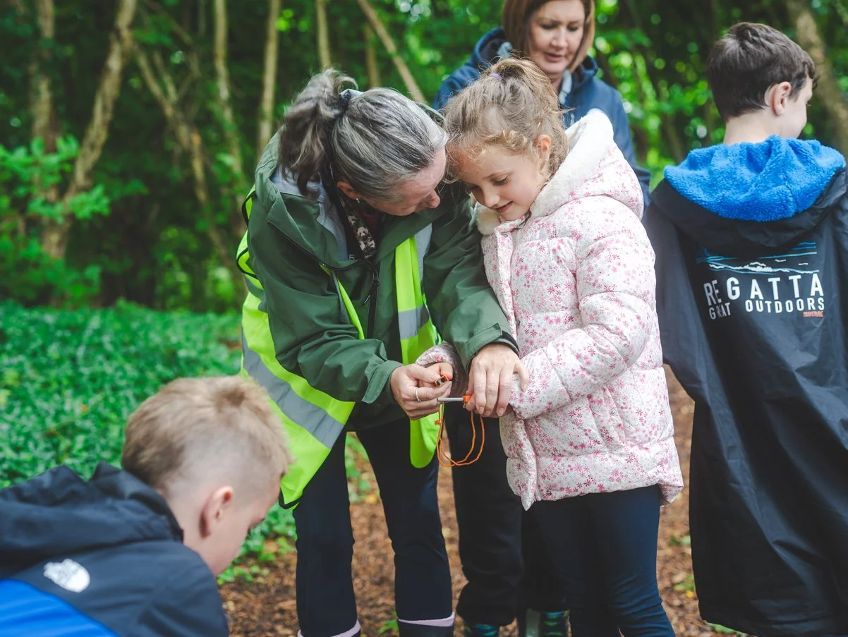 Group of children and an adult woman in a forest, participating in an outdoor educational activity, with one girl and woman focusing on a small object in their hands.