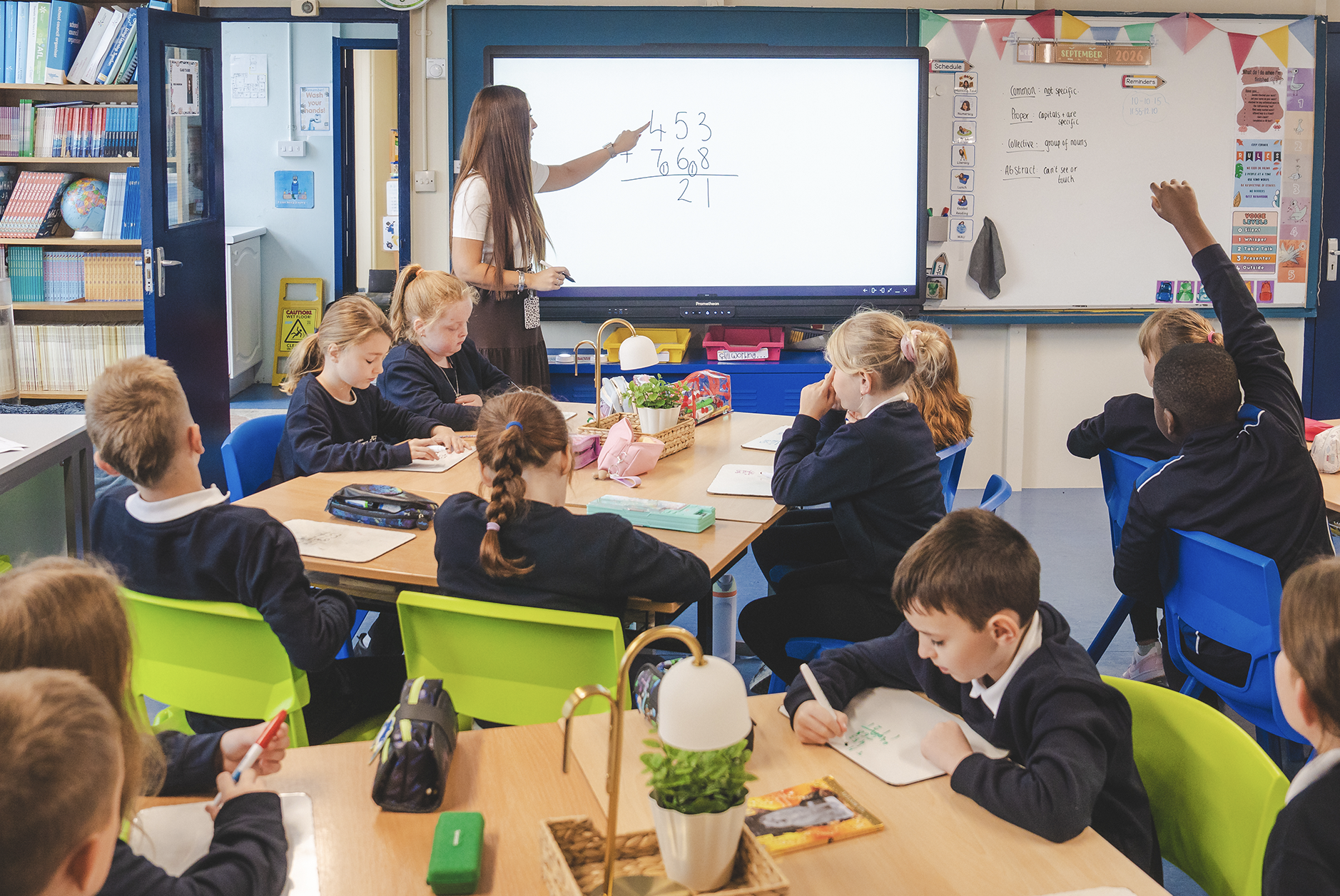 A classroom scene with students sitting at tables, some with raised hands, listening to a teacher at the front writing math problems on an interactive whiteboard.
