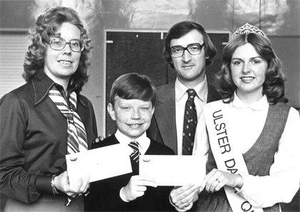 A group of four people, two women, one man, and a girl, standing indoors. The girl wears a sash and crown, holding a large check, suggesting a pageant or contest award. The woman on the left is in glasses and a striped tie, possibly an organizer or teacher.