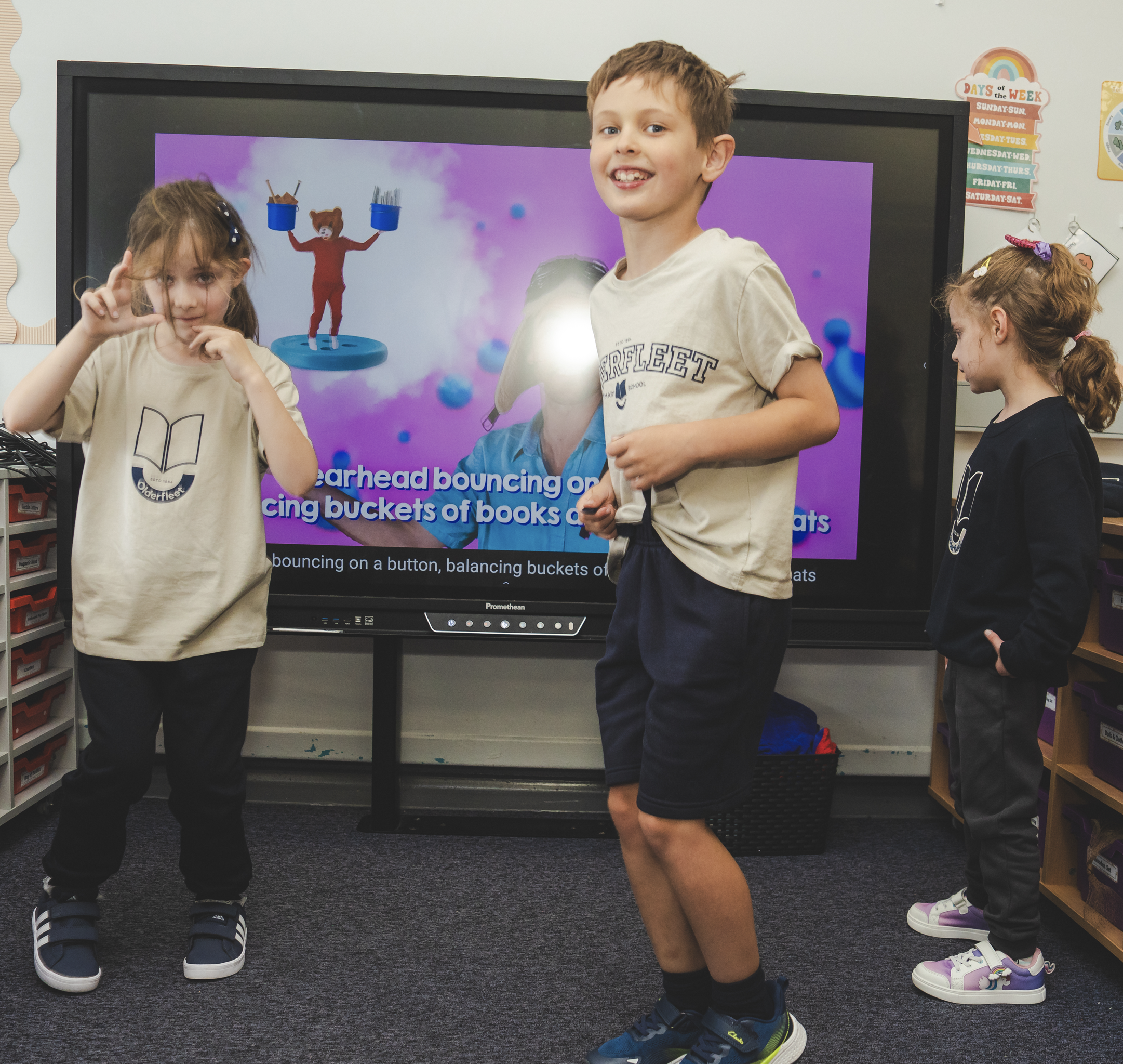 Children standing in a classroom, with a digital screen displaying a bouncing bear mascot and text about bouncing on a button, balancing buckets of books and snacks. Kids wear casual clothing and sneakers.