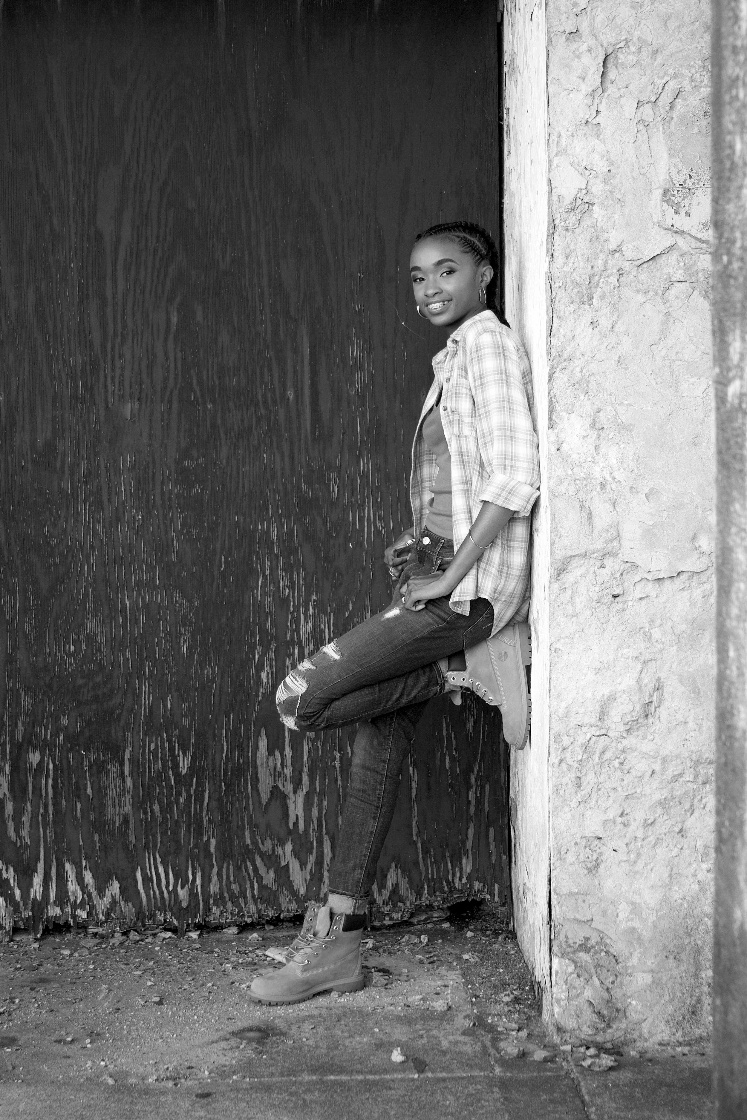A young woman leaning against a wall, smiling at the camera, wearing ripped jeans, a plaid shirt, and boots, standing outdoors.
