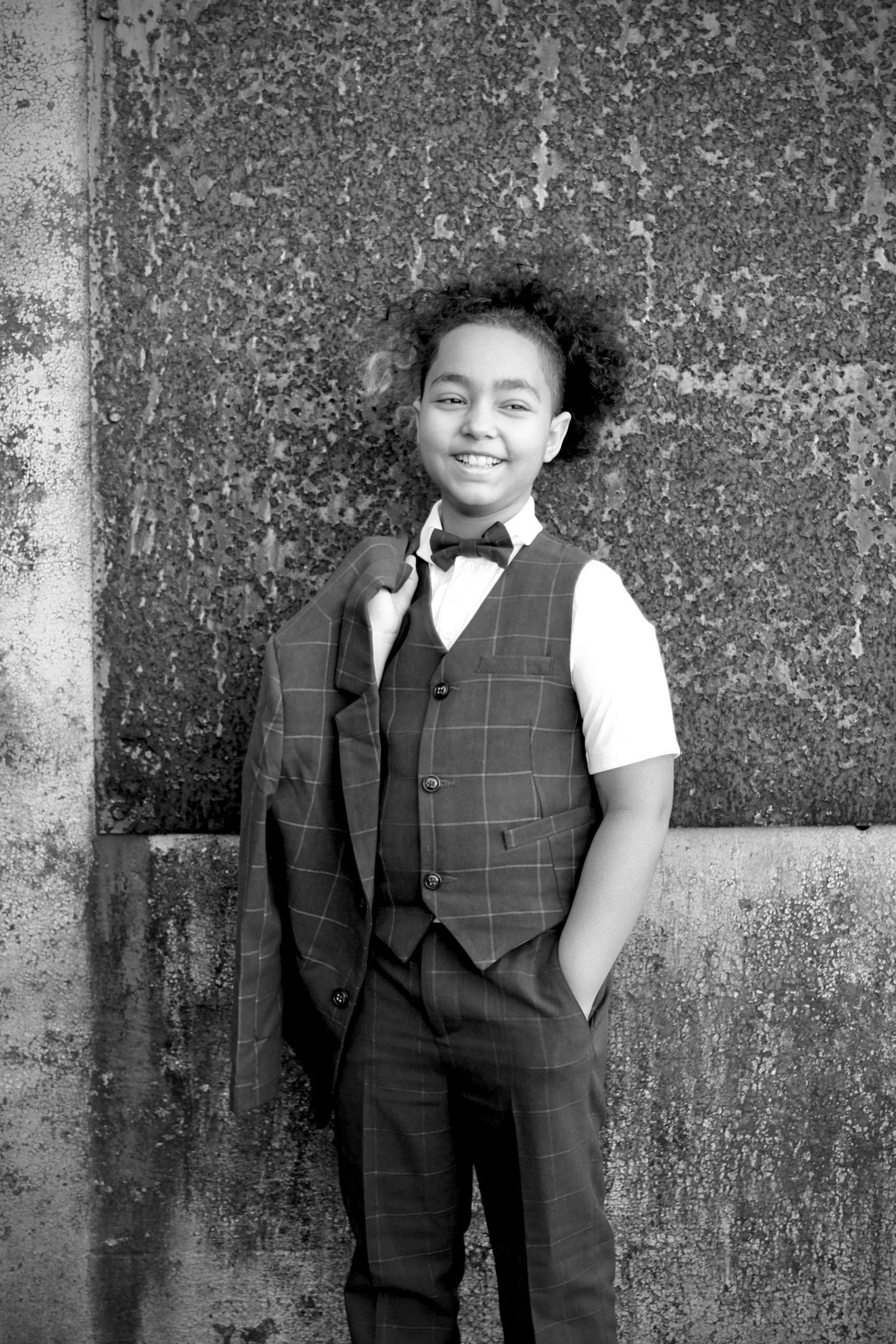 A young girl dressed in a suit with a bow tie, holding a jacket over her shoulder, standing against a textured wall, smiling in a black and white photo.