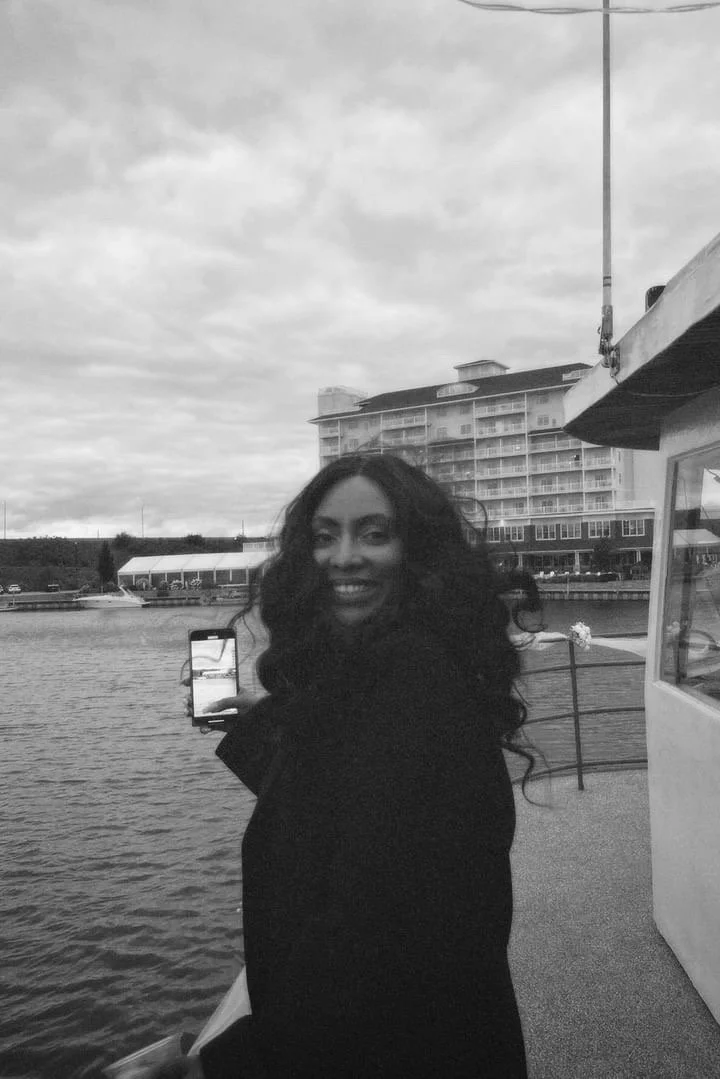 A woman with long, curly hair smiling and holding a smartphone on a boat near a body of water, with a building and cloudy sky in the background.