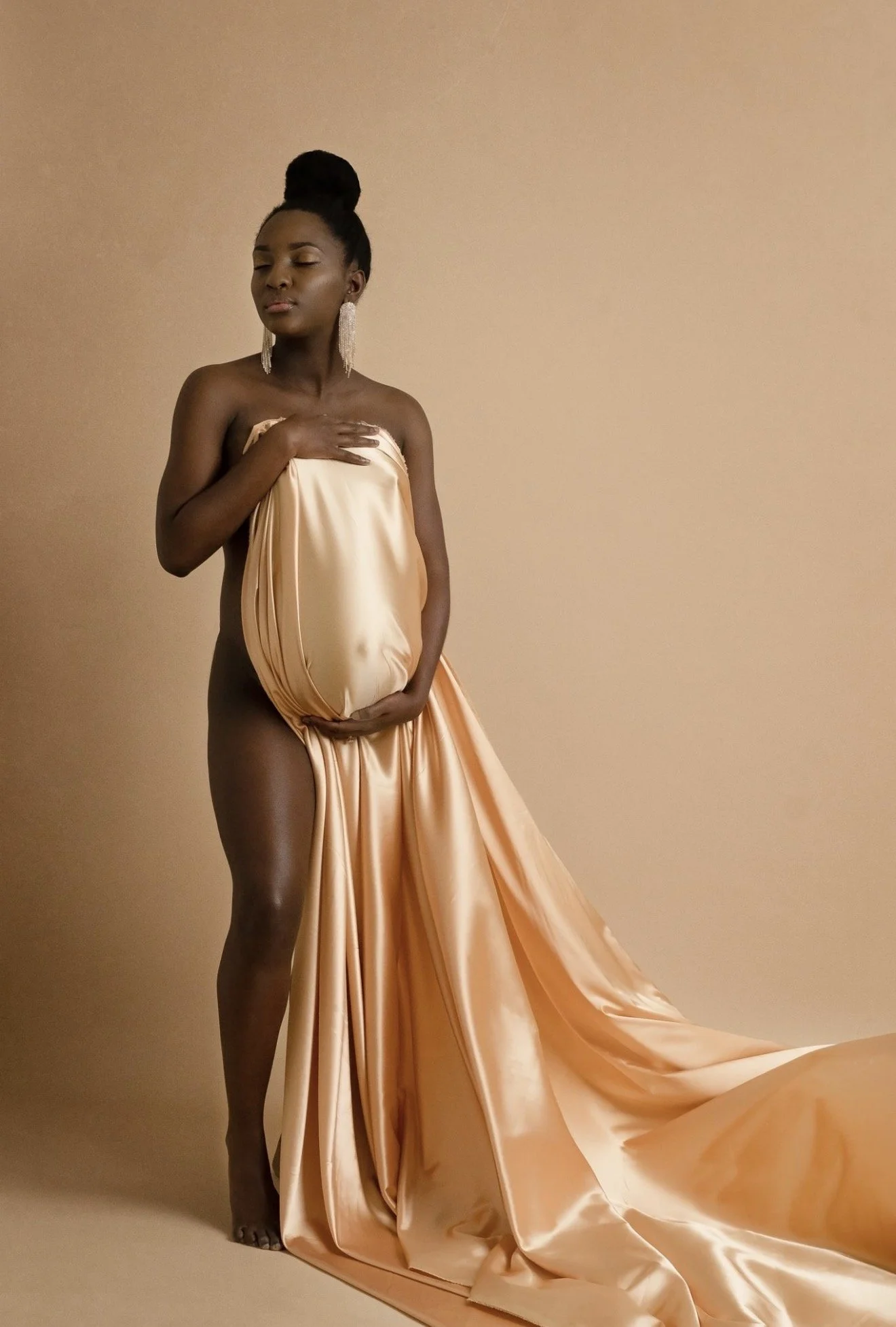 A woman with dark skin and natural hair styled up, standing against a plain beige background. She is topless, covering her chest with her right hand, and is holding a large piece of peach-colored satin fabric that drapes from her waist to the floor.