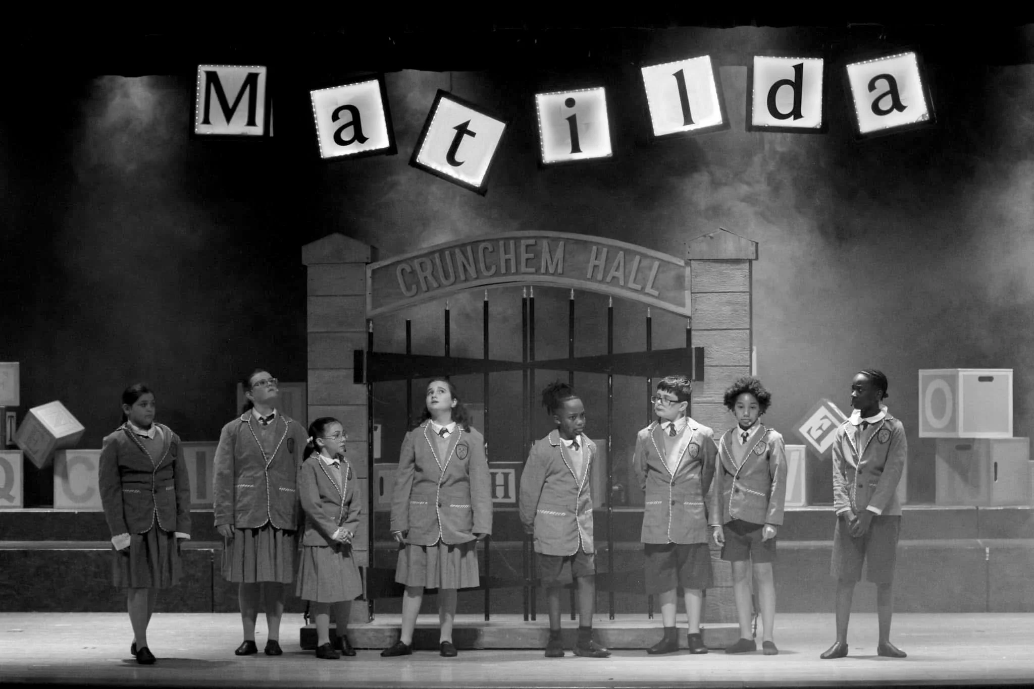 Black and white photo of children in school uniforms standing on stage in front of a backdrop with the words 'CRUNCHEM HALL' and decor including large alphabet blocks and the name 'Matilda' spelled out above.