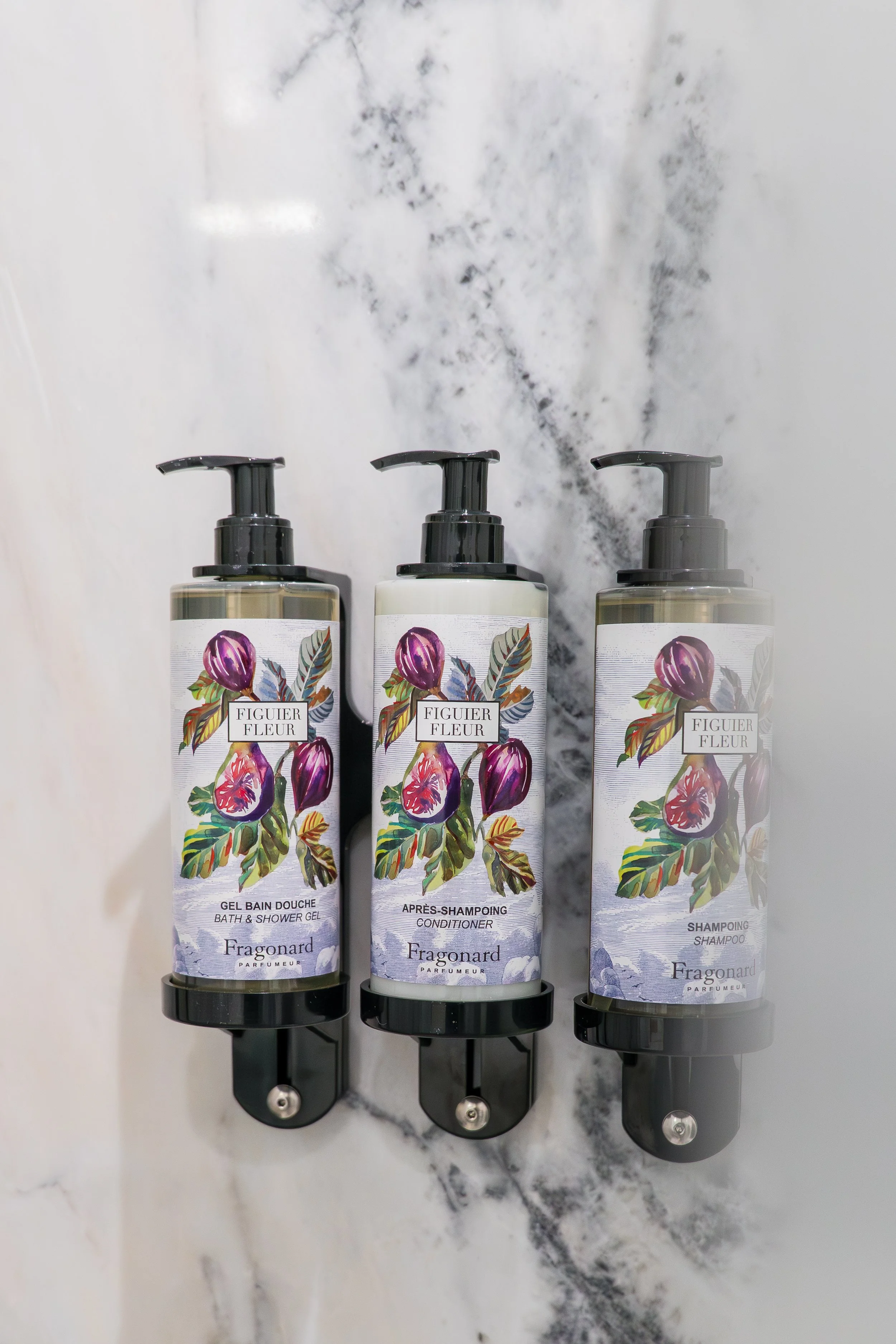 Three bottles of Fragonard fig tree floral scent products on a black metal shelf on a marble countertop. The bottles are labeled as bath and shower gel, conditioner, and shampoo.