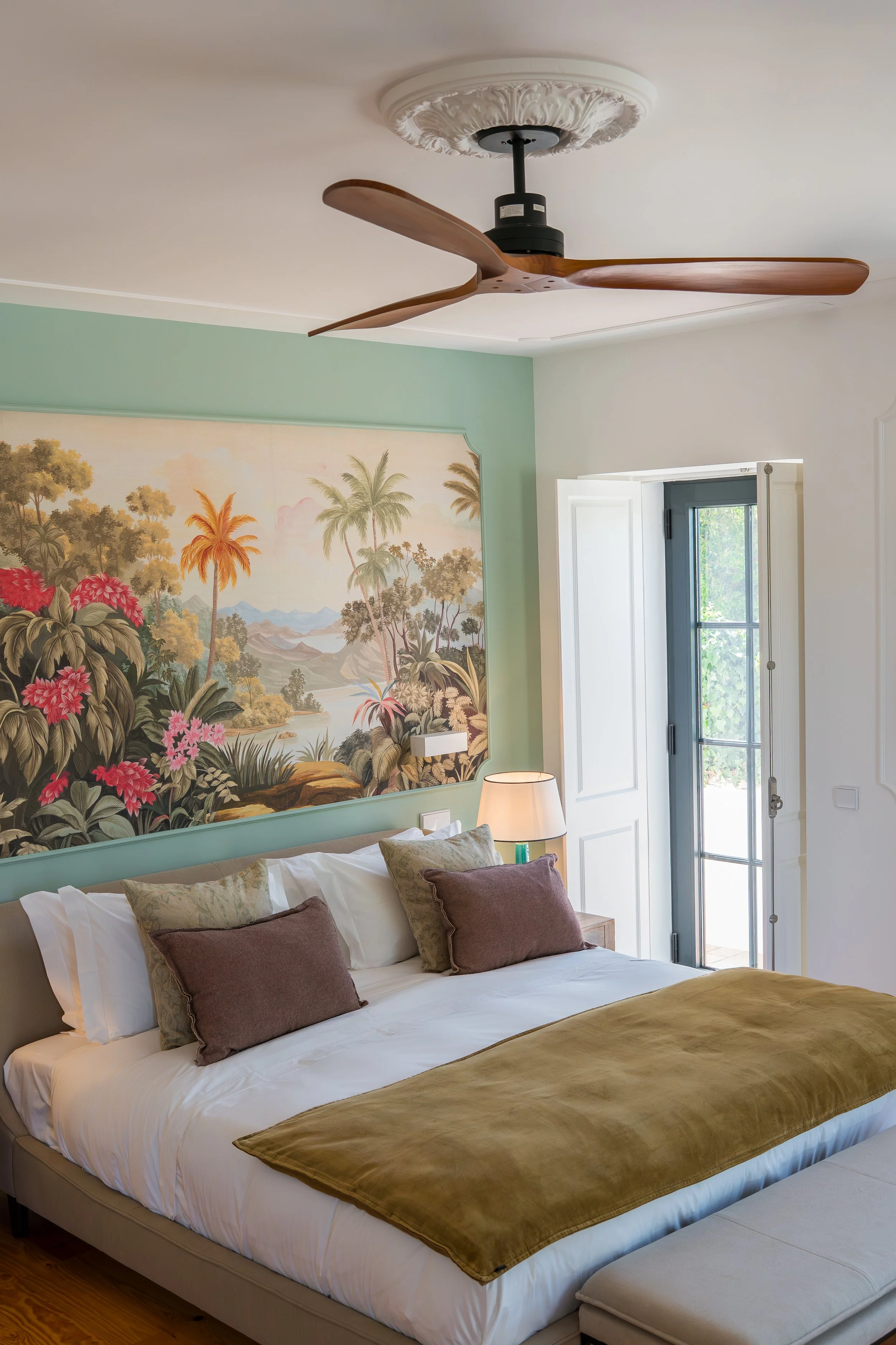 A bedroom with a large bed with white sheets, multiple pillows, and a brown blanket. A wall mural depicting a tropical landscape with palm trees and mountains. A ceiling fan with wooden blades. A window with white shutters and a door leading outside.