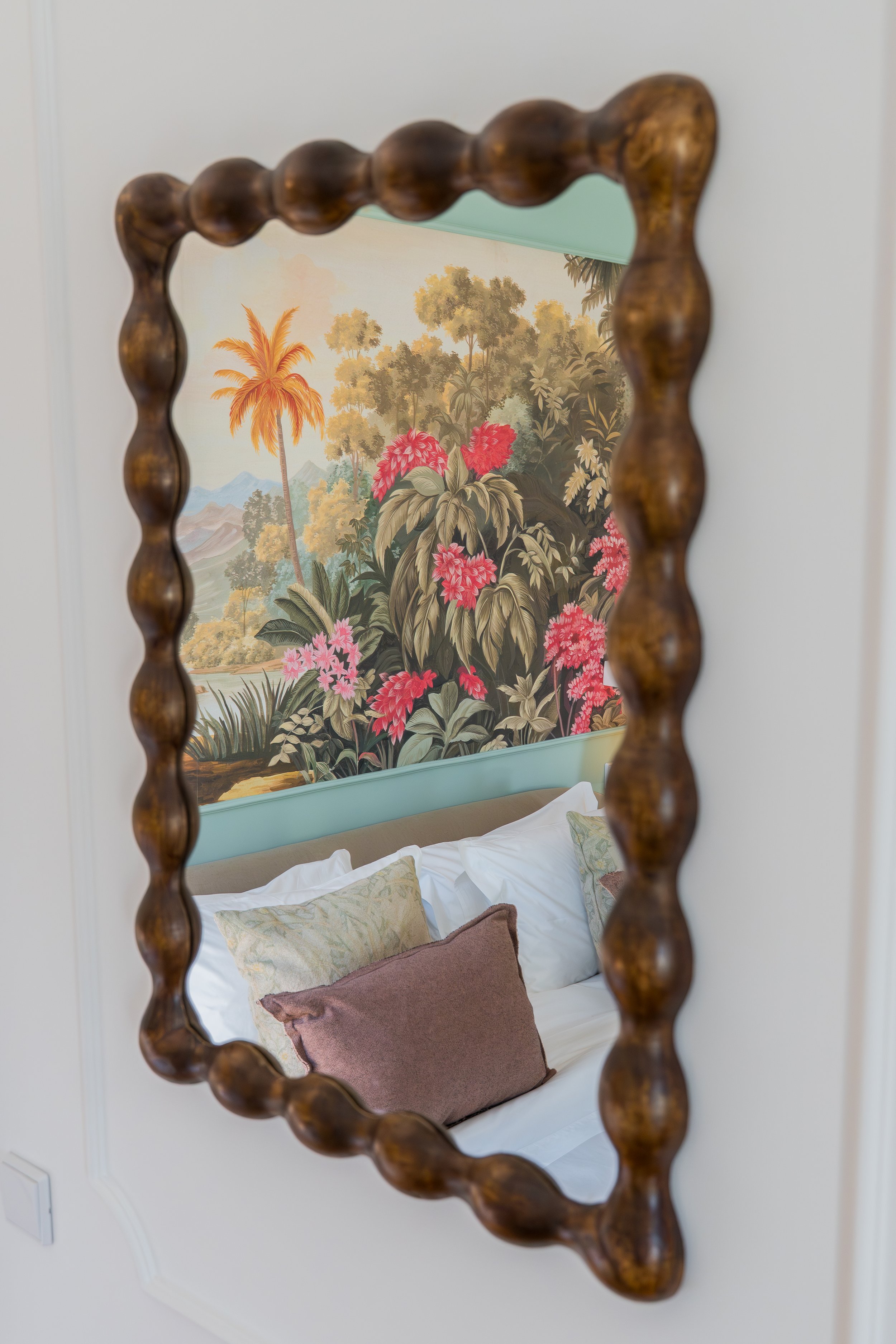 Decorative mirror with a wooden frame reflecting a bed with pillows and a tropical rainforest mural.