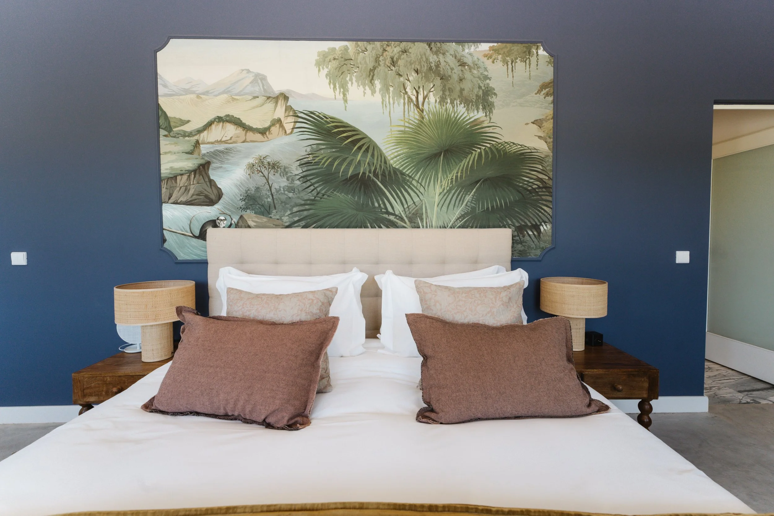 A bedroom with dark blue walls featuring a large painted mural of lush greenery, trees, and mountains, with a river flowing through the landscape. The bed has a beige upholstered headboard, white bedding, and multiple brown and beige pillows. There a