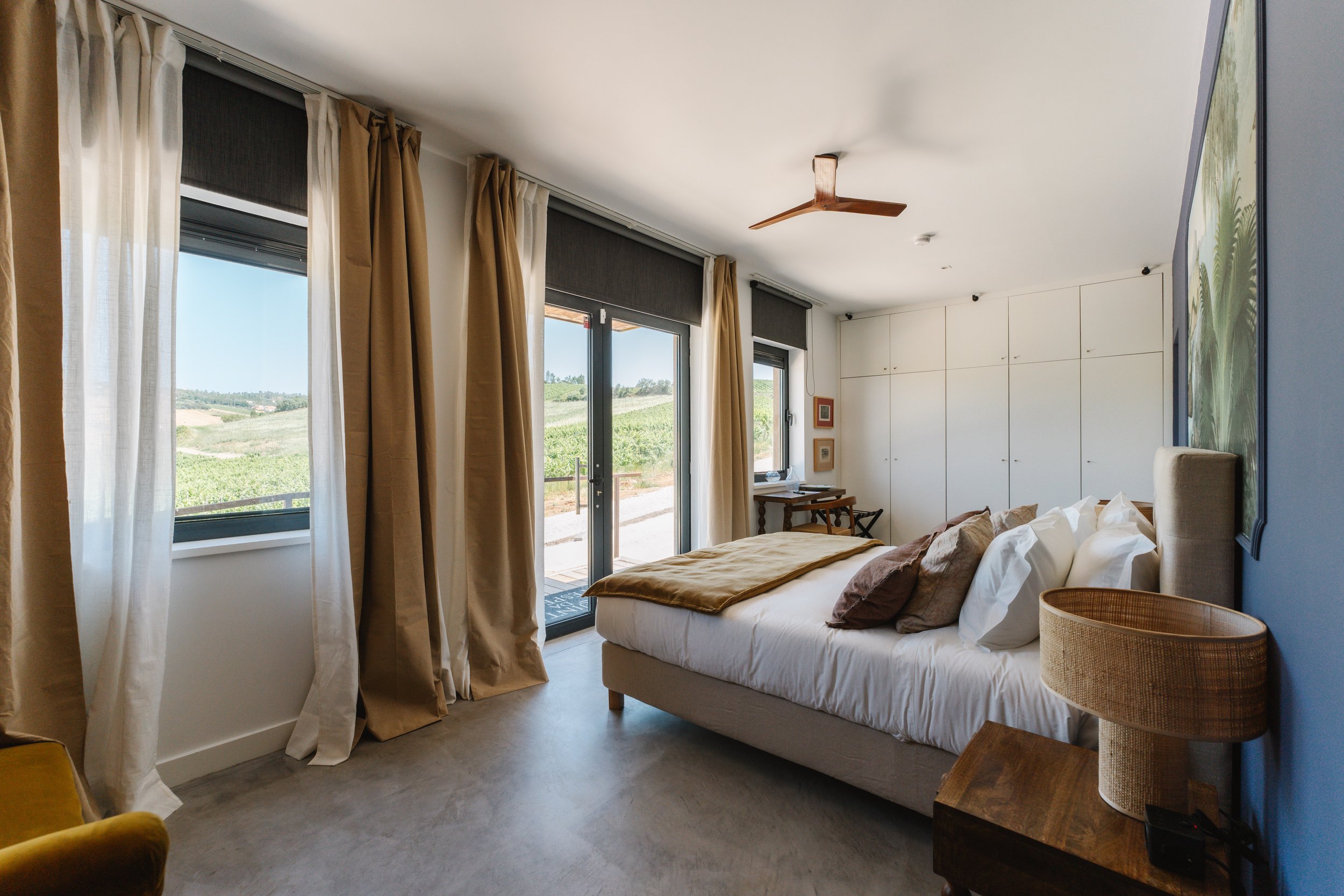 A cozy bedroom with a large bed, beige and white bedding, and a small bedside table with a woven lamp. Large windows with beige and white curtains show a view of green fields outdoors. A ceiling fan is mounted on the white ceiling, and the room has m