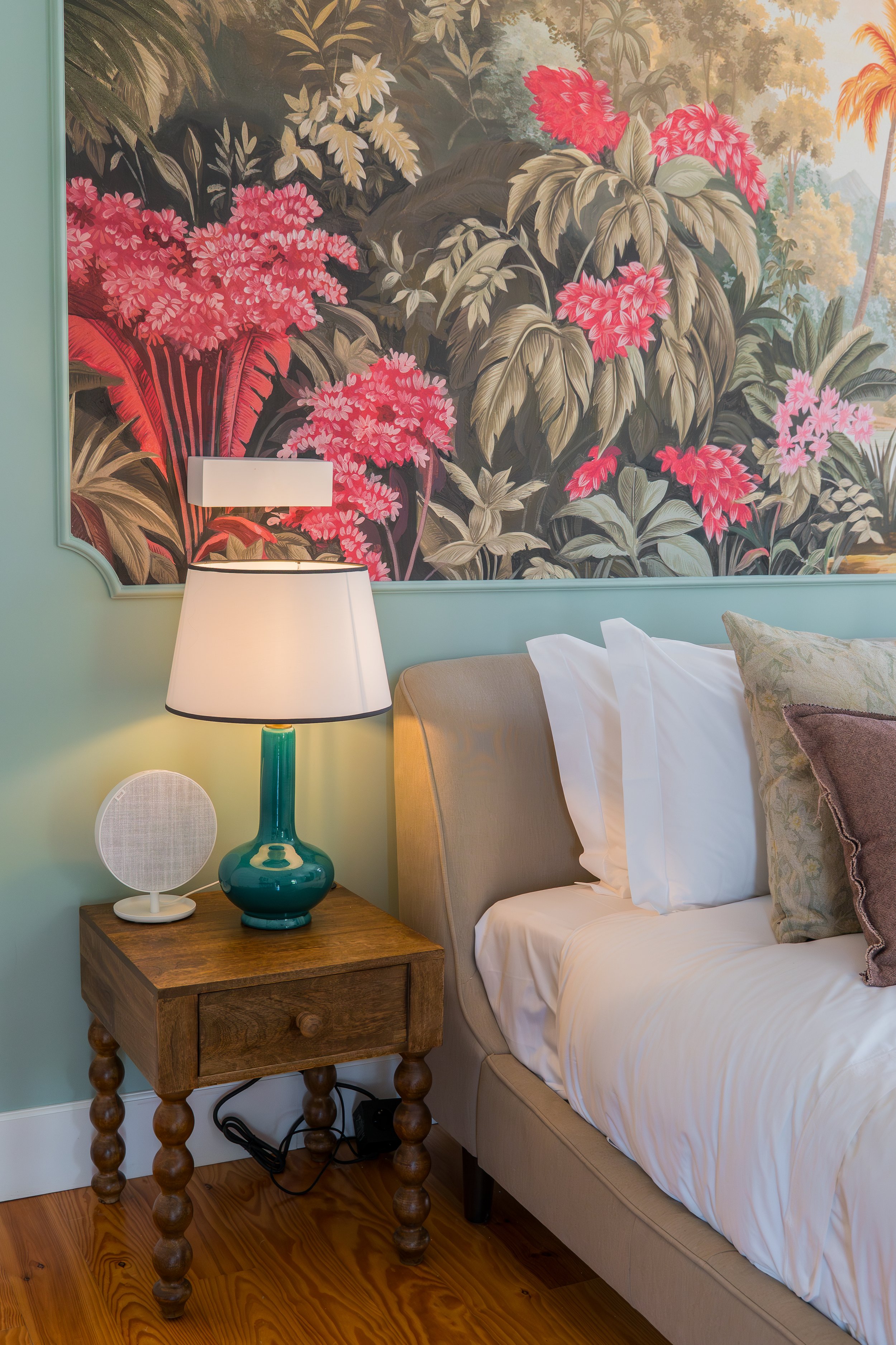 A bedroom with a beige bed, white pillows, a wooden nightstand, a green table lamp with a white shade, a small white speaker, and a vibrant botanical mural with pink, red, and beige plants on the wall behind the bed.