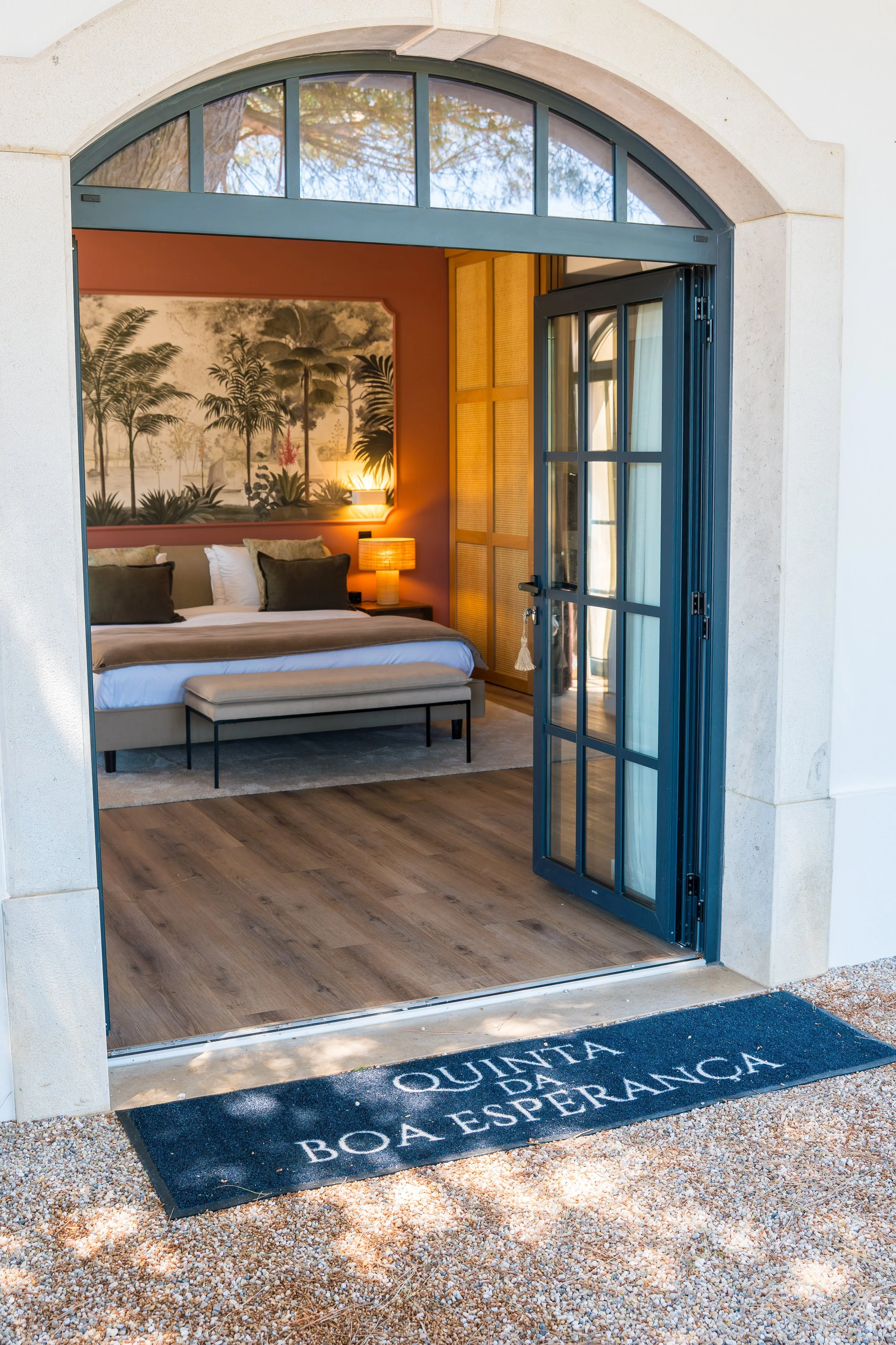 Open doorway leading into a bedroom with a bed, decorative wall art, and warm lighting, seen from outside on a pebble-covered ground with a black rug reading 'QUINTA ROA ESPERANZA'.