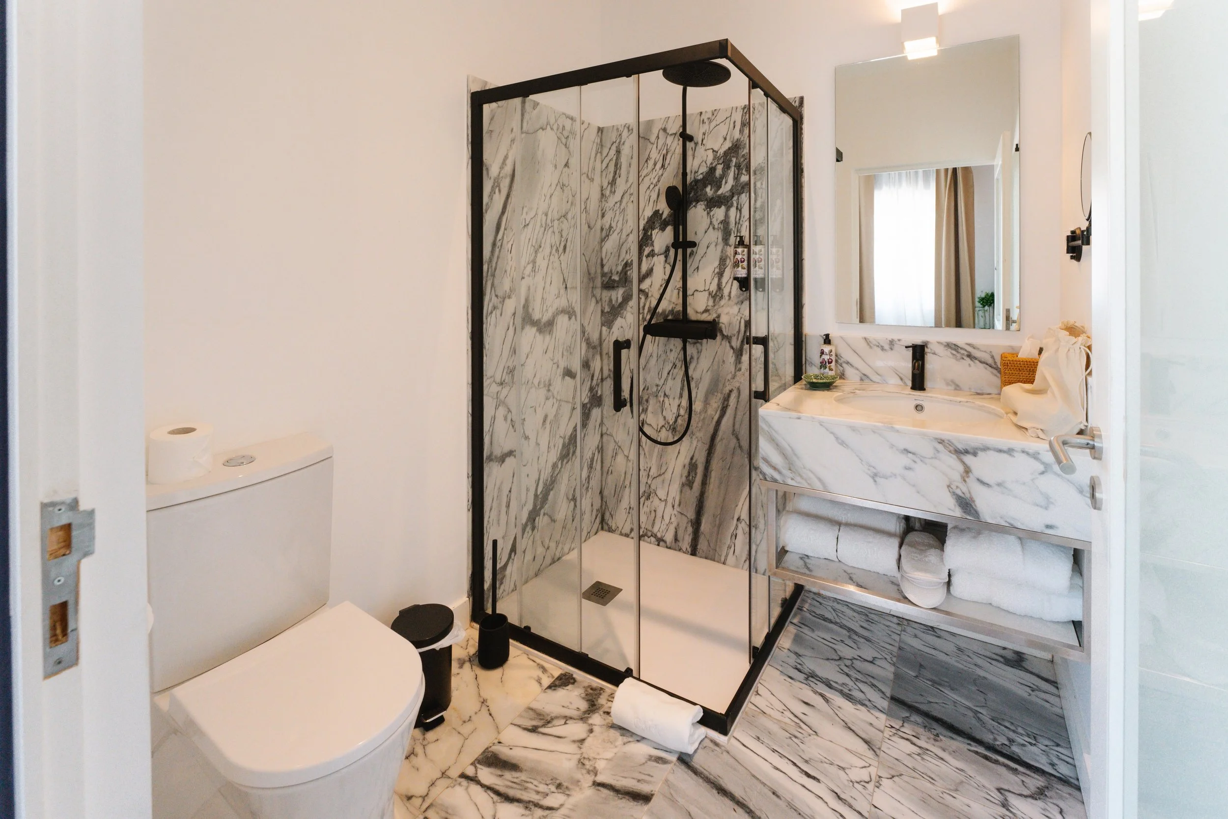 A modern bathroom featuring a glass-enclosed shower with marble walls and a black showerhead, a marble vanity with a large mirror, a white toilet, and matching marble flooring. Towels are stored on a shelf below the vanity, and decorative items are o