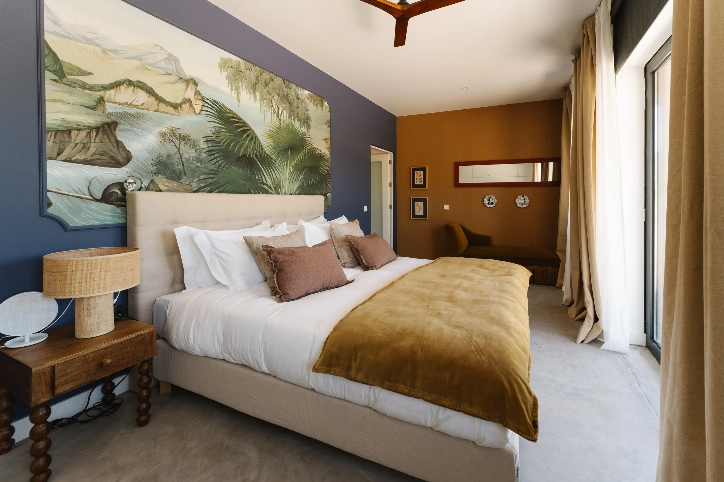 A bedroom with a large bed with white sheets and brown pillows, a wooden bedside table with a beige lamp, a colorful wall mural of a jungle scene behind the bed, and a seating area with curtains and framed pictures on the wall.