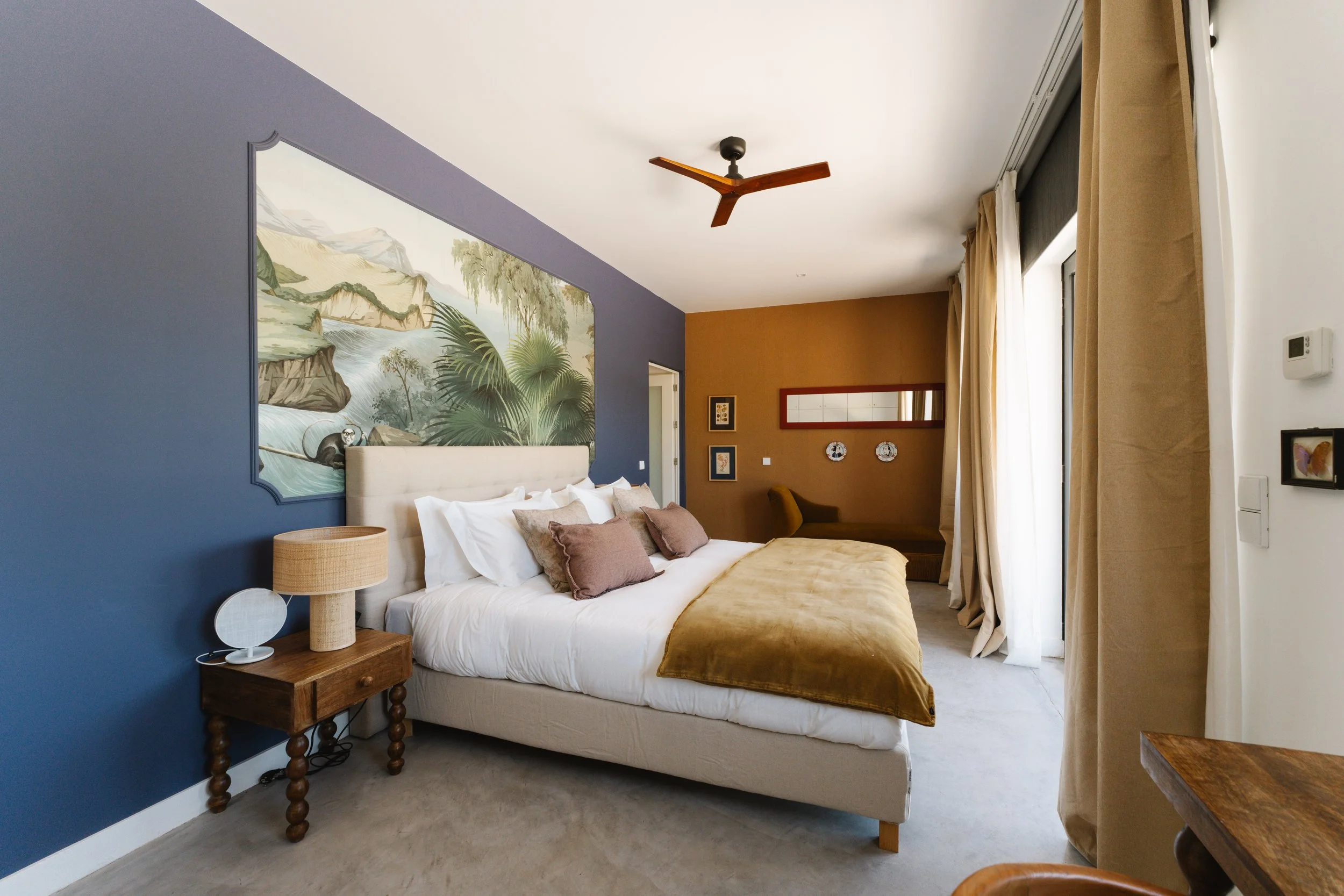 A bedroom with a white bed, beige bedding, and multiple pillows, against a blue and mustard wall, with artwork and windows with beige curtains.