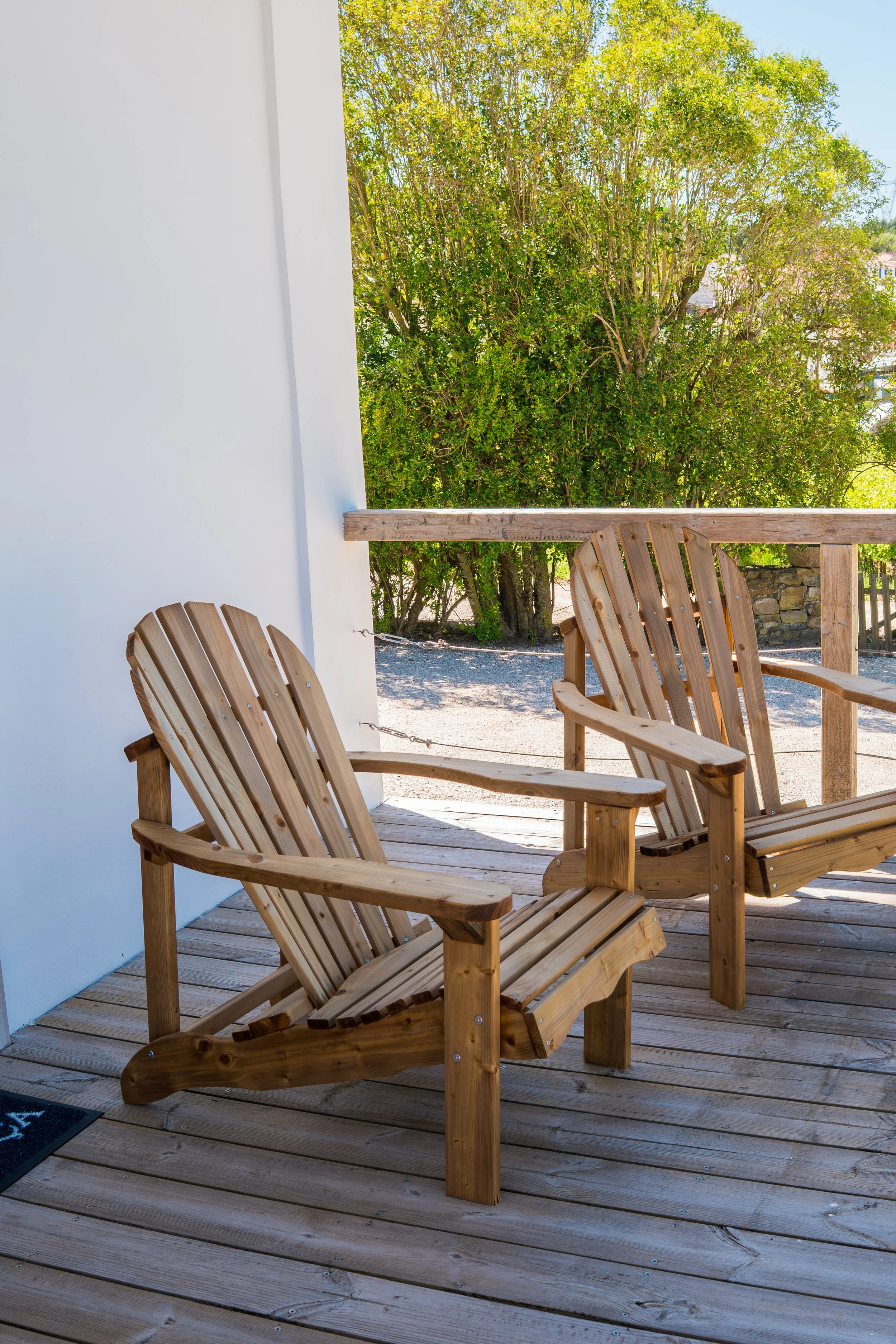 Wooden outdoor deck with two Adirondack chairs and a railing, overlooking trees and a clear blue sky.