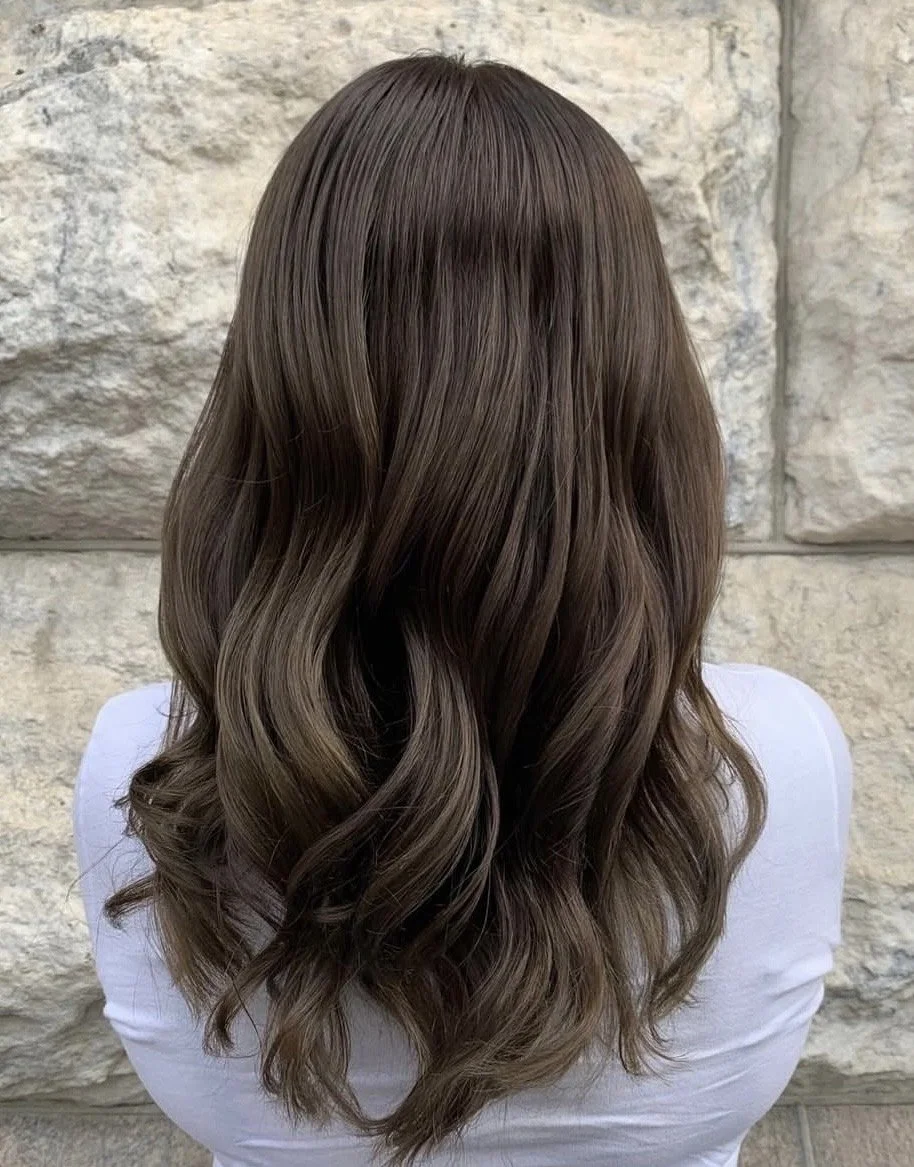 Back view of a person with long wavy brown hair standing against a stone wall.