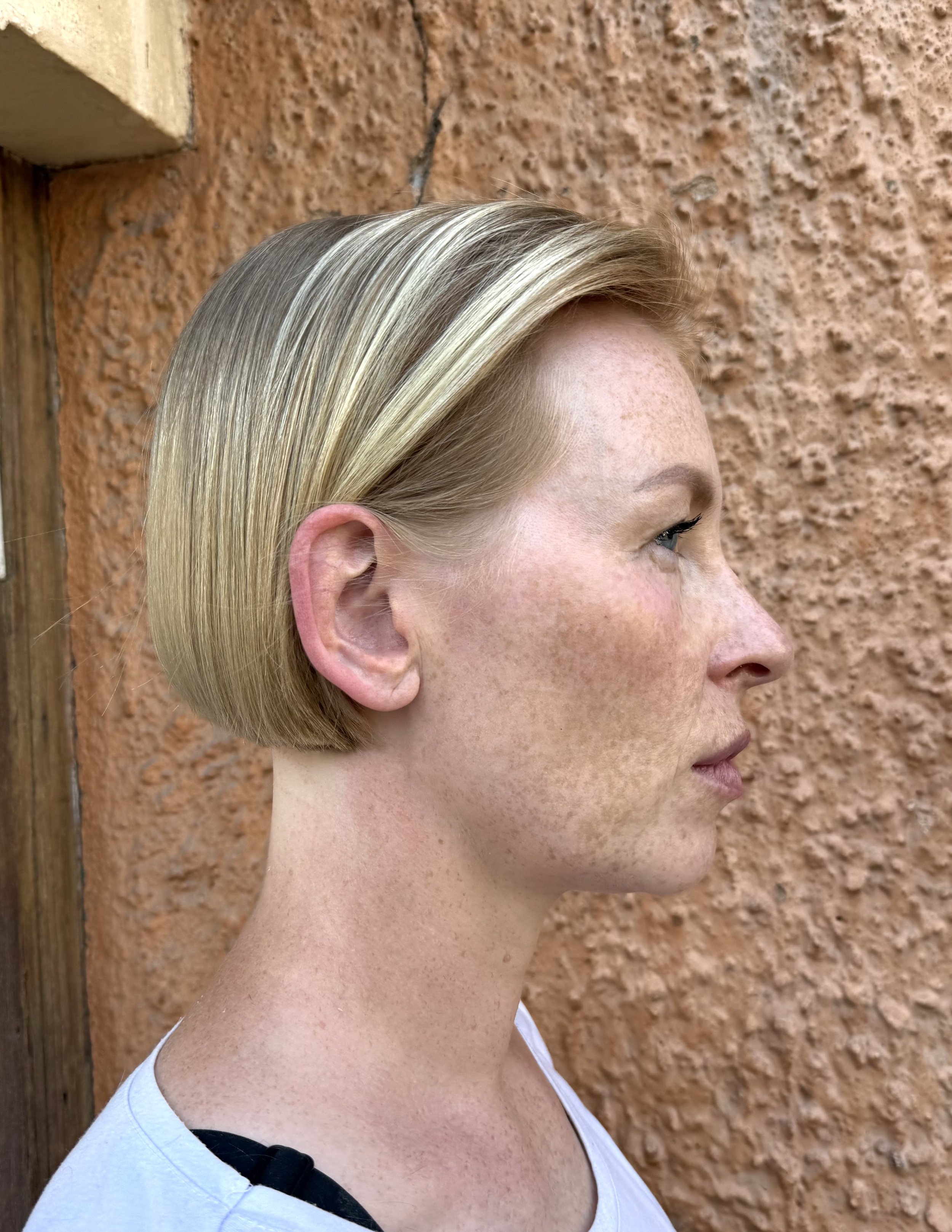 Side profile of a woman with short, blonde hair standing against a textured, orange-brown wall.