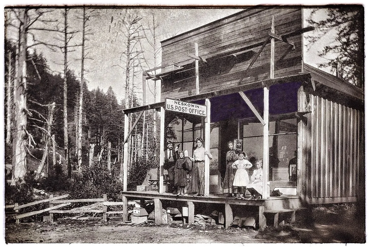 A black and white photograph of a rustic wooden building with a sign that reads 'Neskowin U.S. Post Office.' Several people, including women and children, are standing on the porch, with some sitting and others standing, holding packages and conversing. The background features tall, leafless trees and a forested area.