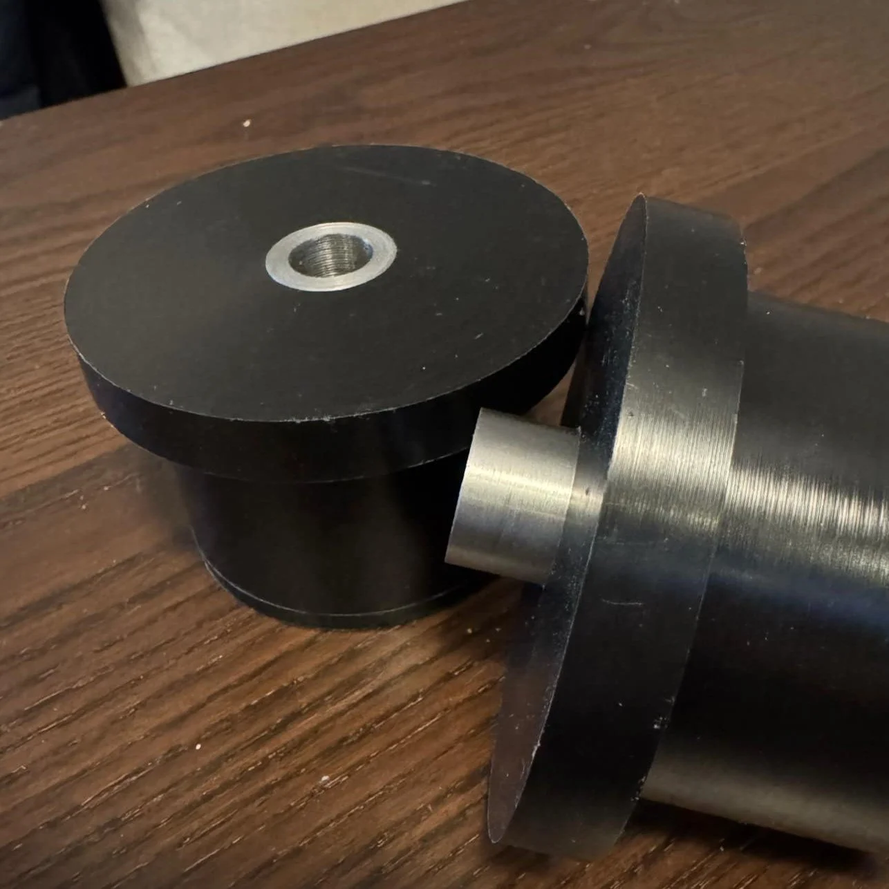 Close-up photo of a metal roller with a threaded hole in the center, attached to a larger cylindrical metal part, placed on a wooden surface.