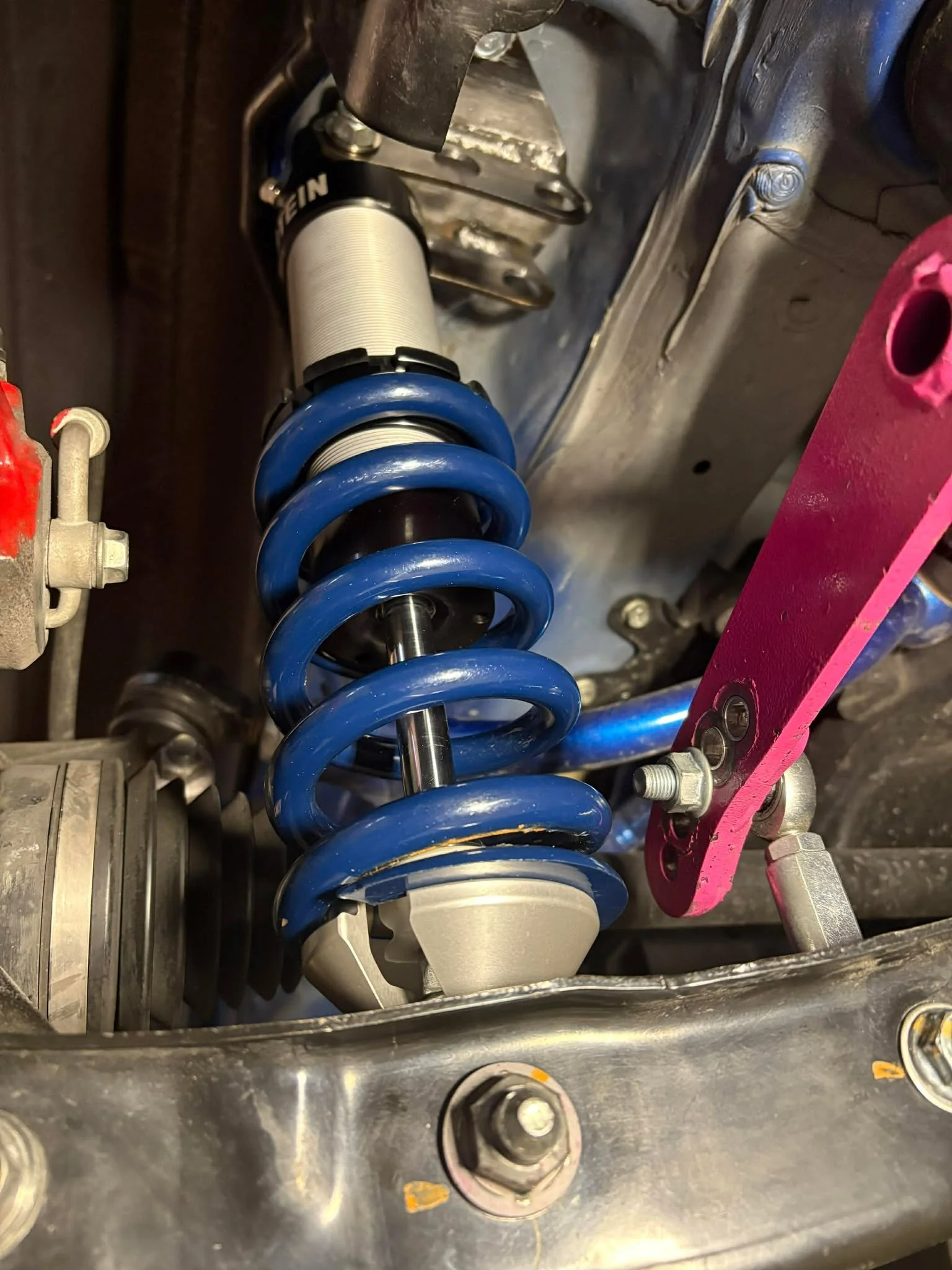 Close-up view of a vehicle's suspension coil spring and shock absorber assembly mounted on a car's chassis, with various bolts and metal components visible.