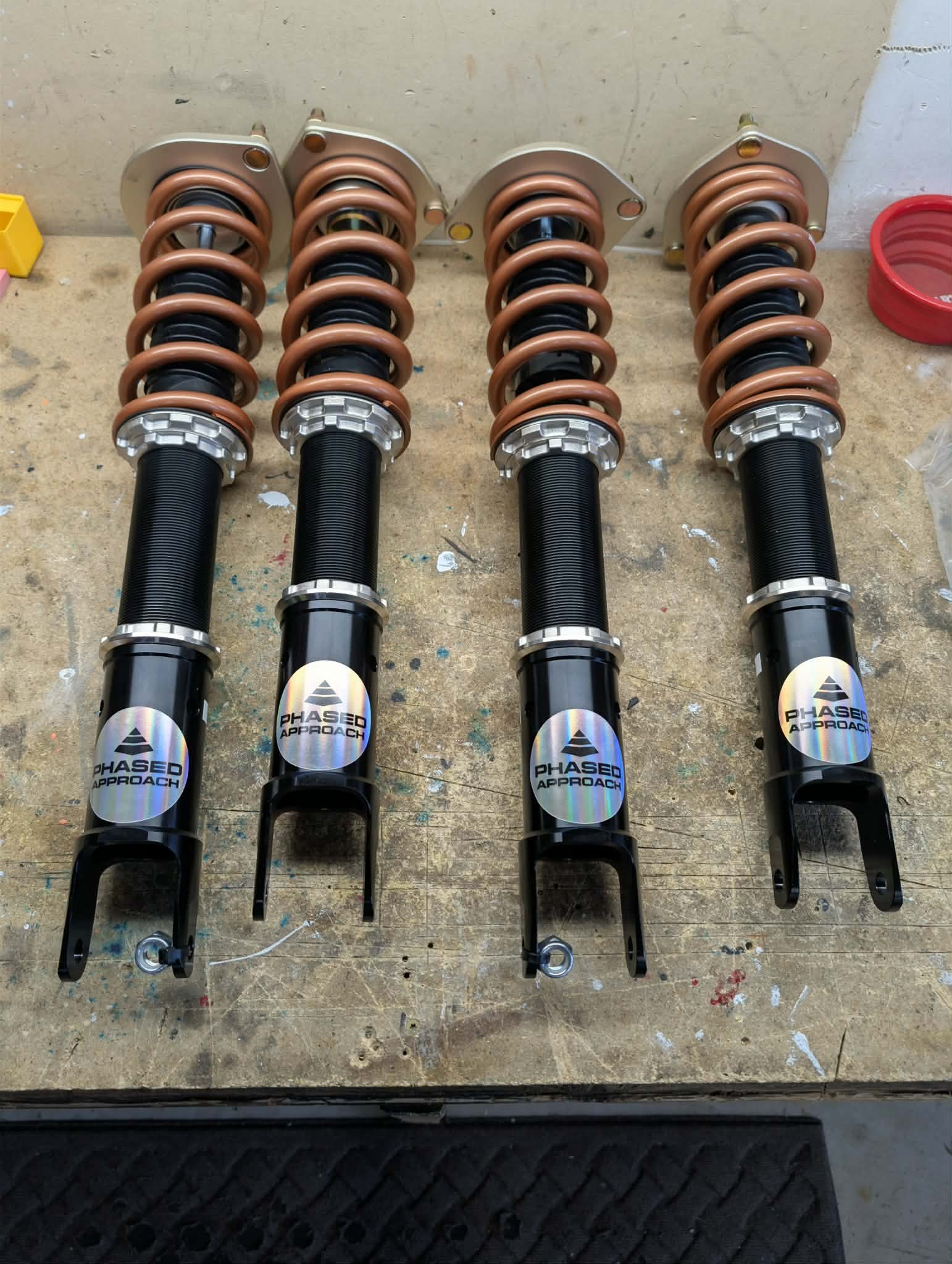 Four black coilover suspension struts with copper-colored springs and black mounting brackets placed on a wooden workbench.