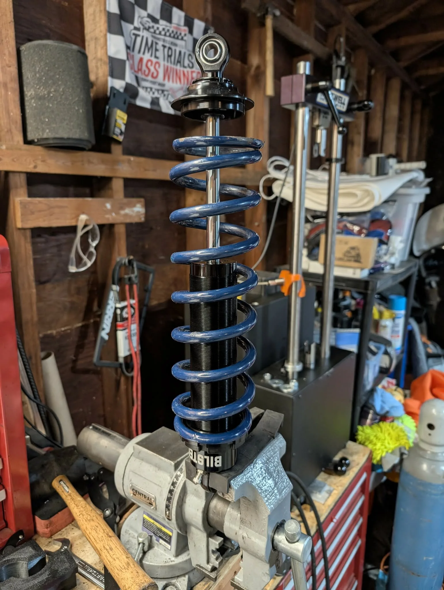 A shock absorber mounted on a workbench in a garage or workshop, with clamps holding it in place. The background shows tools, a poster, and various workshop items.