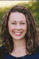 A woman with curly brown hair smiling outdoors against a blurred background of trees and greenery.  This is Krystal Saxey, our billing and credentialling specialist.