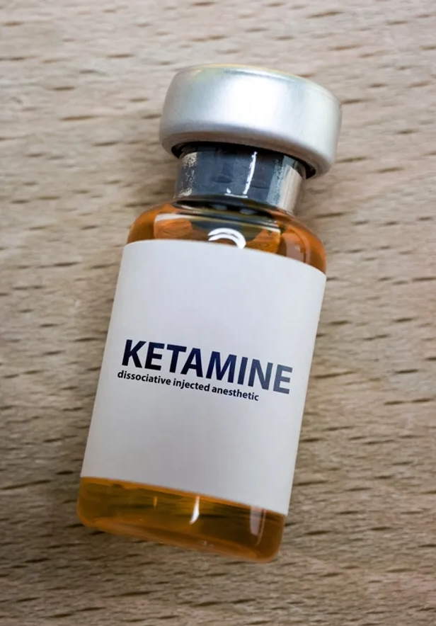 A small glass vial with an orange liquid, labeled 'KETAMINE' and 'dissociative injected anesthetic,' lying on a wooden surface.  We offer ketamine treatment for those who have treatment resistant depression and those with indicated disorders.