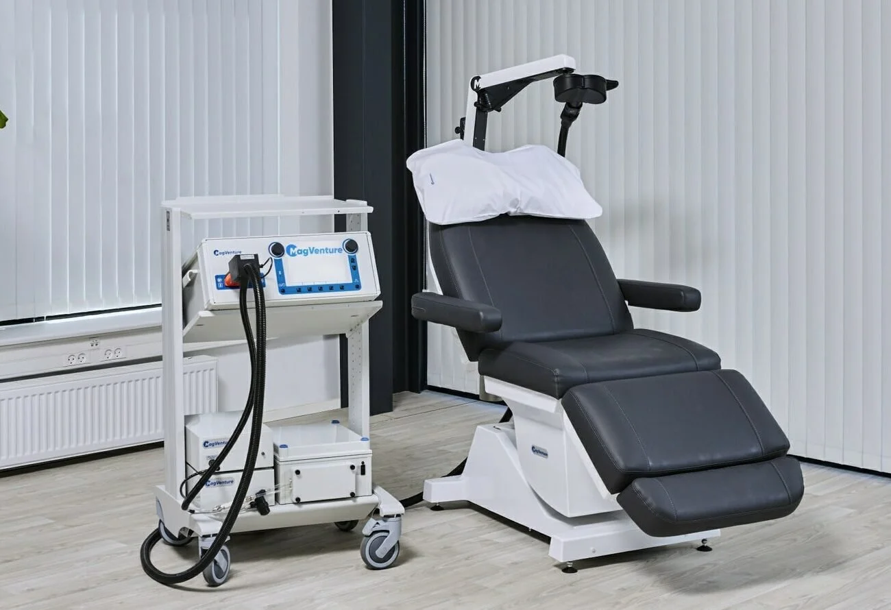Medical examination chair next to a white medical device on a cart, in a clinical room with white vertical blinds.