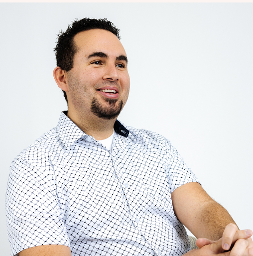 A smiling man with dark hair and a goatee wearing a white shirt with small black checkered pattern, sitting against a plain white background.  This is Ben Saxey PA-C.  He has an MPAS degree and CAQ in psychiatry.