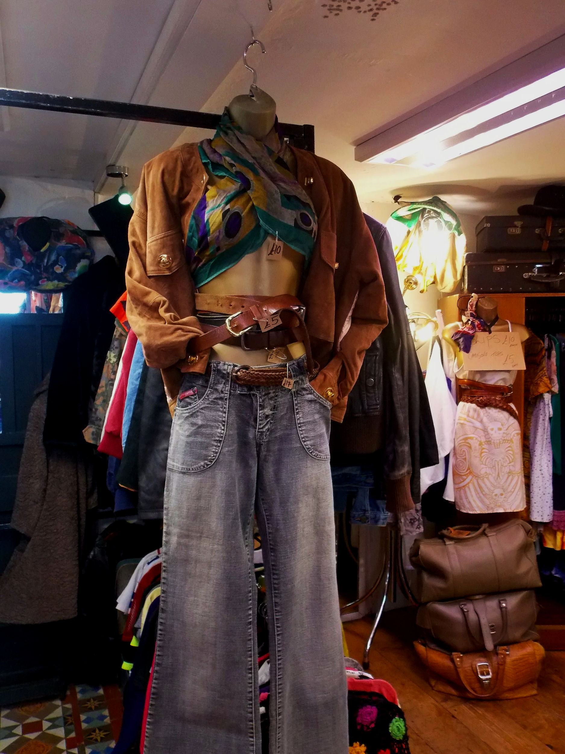 Vintage clothing store display featuring a mannequin dressed in a colorful scarf, brown jacket, belt, and high-waisted jeans, with various clothes and bags in the background.