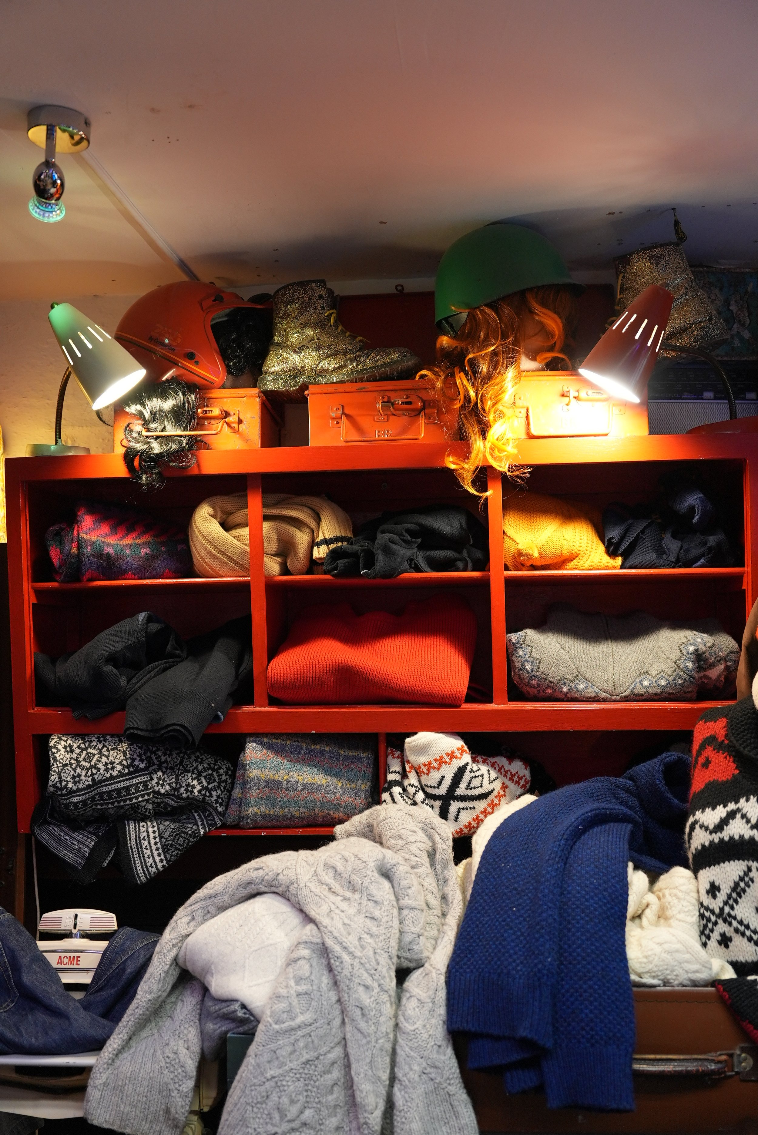 A wooden shelf filled with hats, sweaters, and scarves, with lamps and helmets on top, and piles of clothing in front.