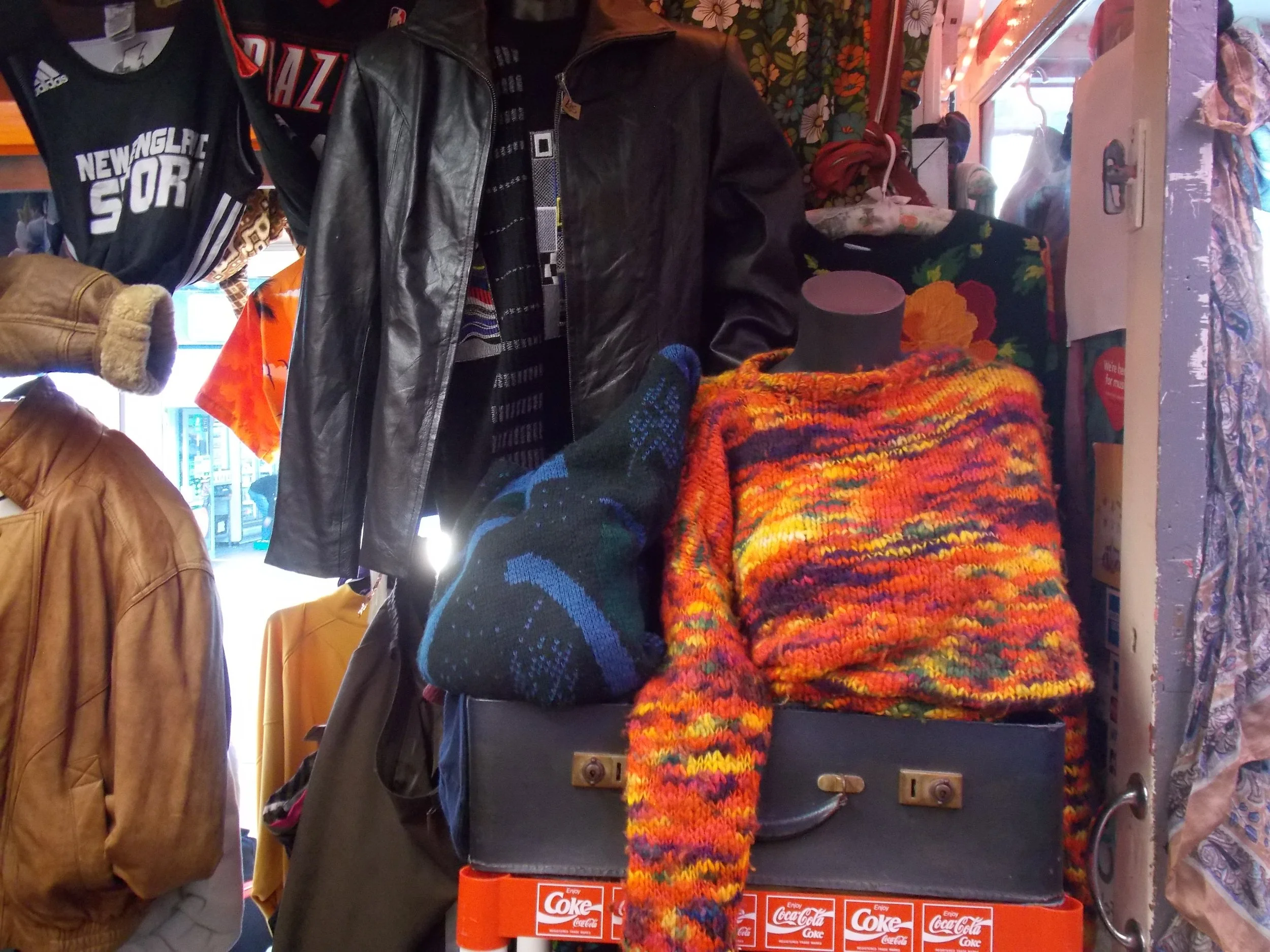 Display of vintage clothing and accessories in a store, including colorful knitted sweaters, leather jackets, and hats.