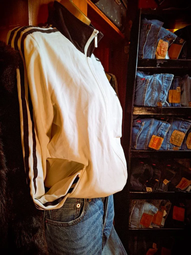 A white zip-up jacket with black stripe details on the sleeves is displayed on a mannequin dressed in blue jeans, in front of a black shelf filled with folded blue jeans.