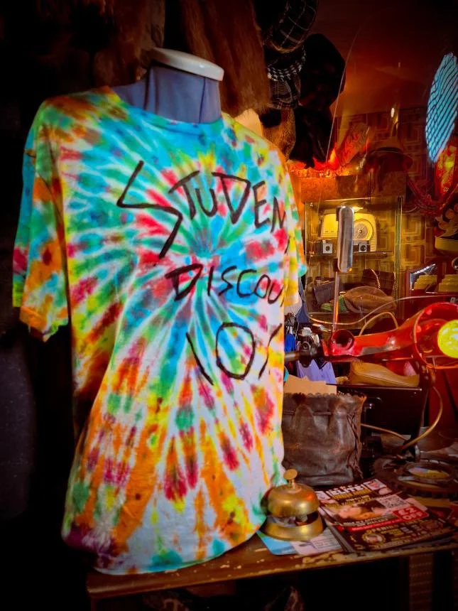 A tie-dye T-shirt on a mannequin with the words 'Student Discount' and a large exclamation point written in black marker.