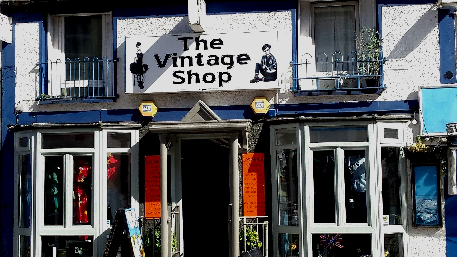 The exterior of a vintage shop named 'The Vintage Shop' with a large sign featuring illustrations of vintage-style women. The shop has bay windows and a small entrance with orange notices on either side.