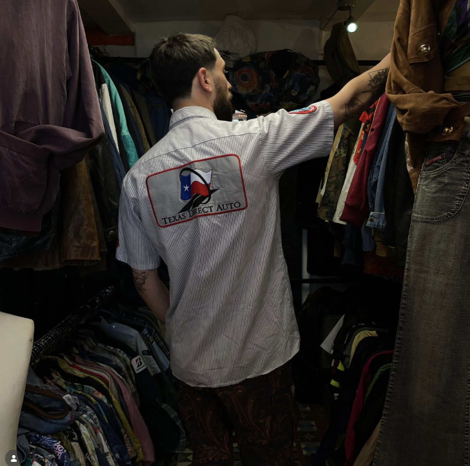 A man wearing a white shirt with the logo of Texas Direct Auto shops for cars. He is shopping for clothes or accessories in a store filled with hanging clothes and backpacks.