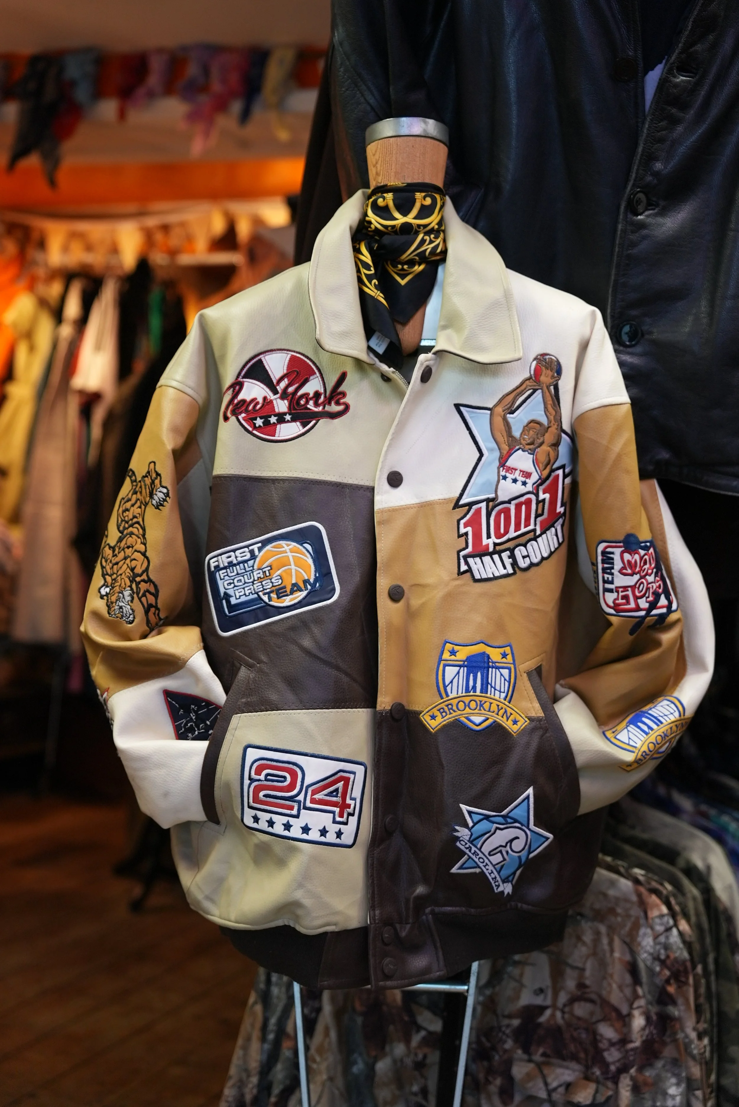 A vintage leather basketball jacket with various patches and embroidered logos, displayed on a mannequin with a black bandana around its neck in a store.