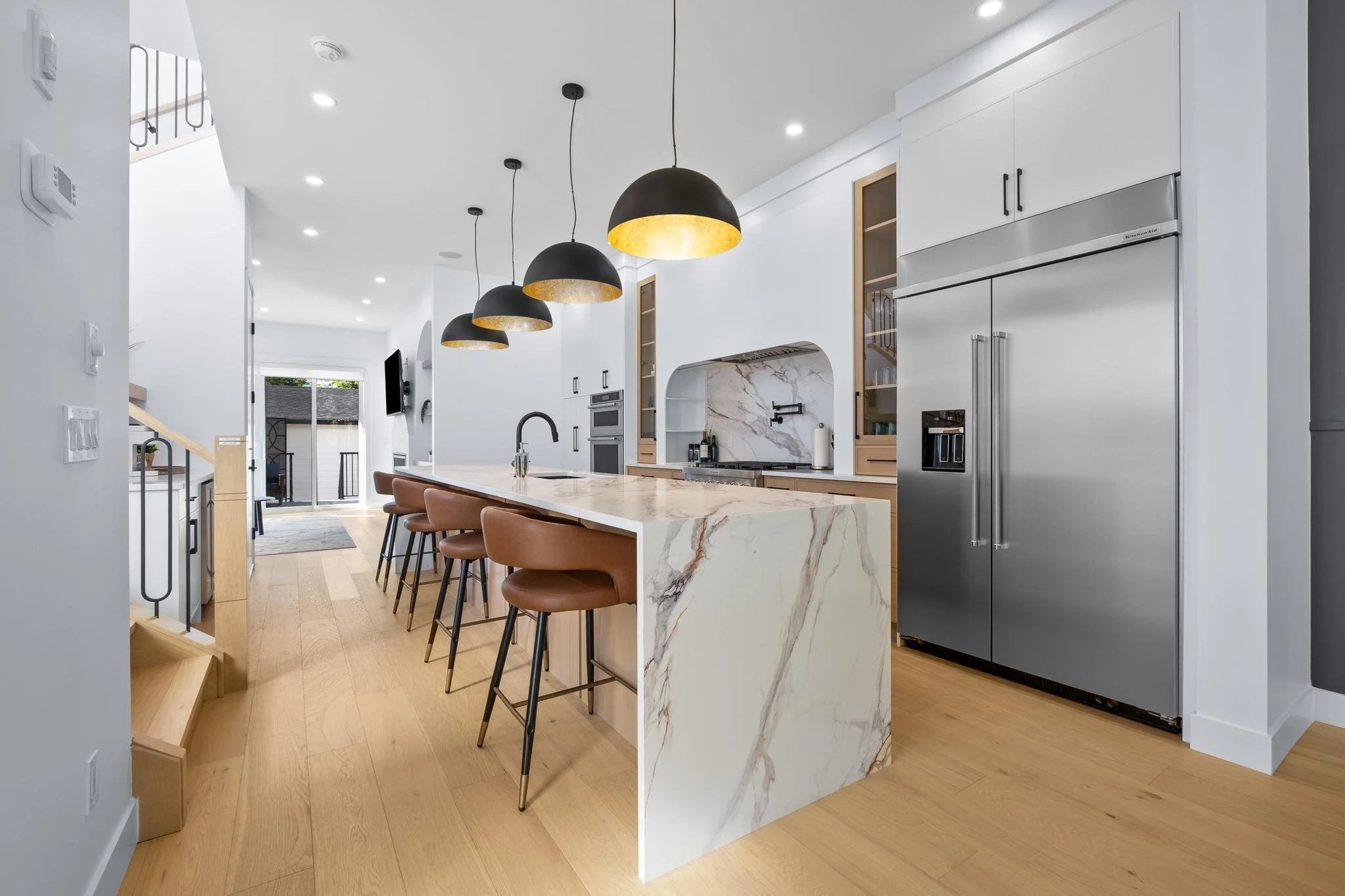 Modern kitchen with white cabinets, marble island, black pendant lights, stainless steel refrigerator, and wooden flooring.