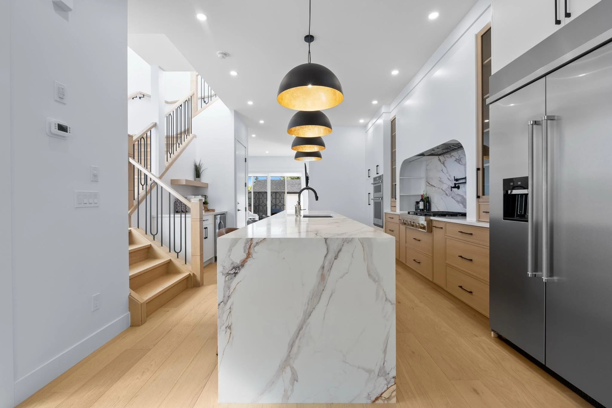Modern kitchen with white walls, wooden flooring, a marble island, wooden cabinets, stainless steel appliances, and a staircase with black railings.