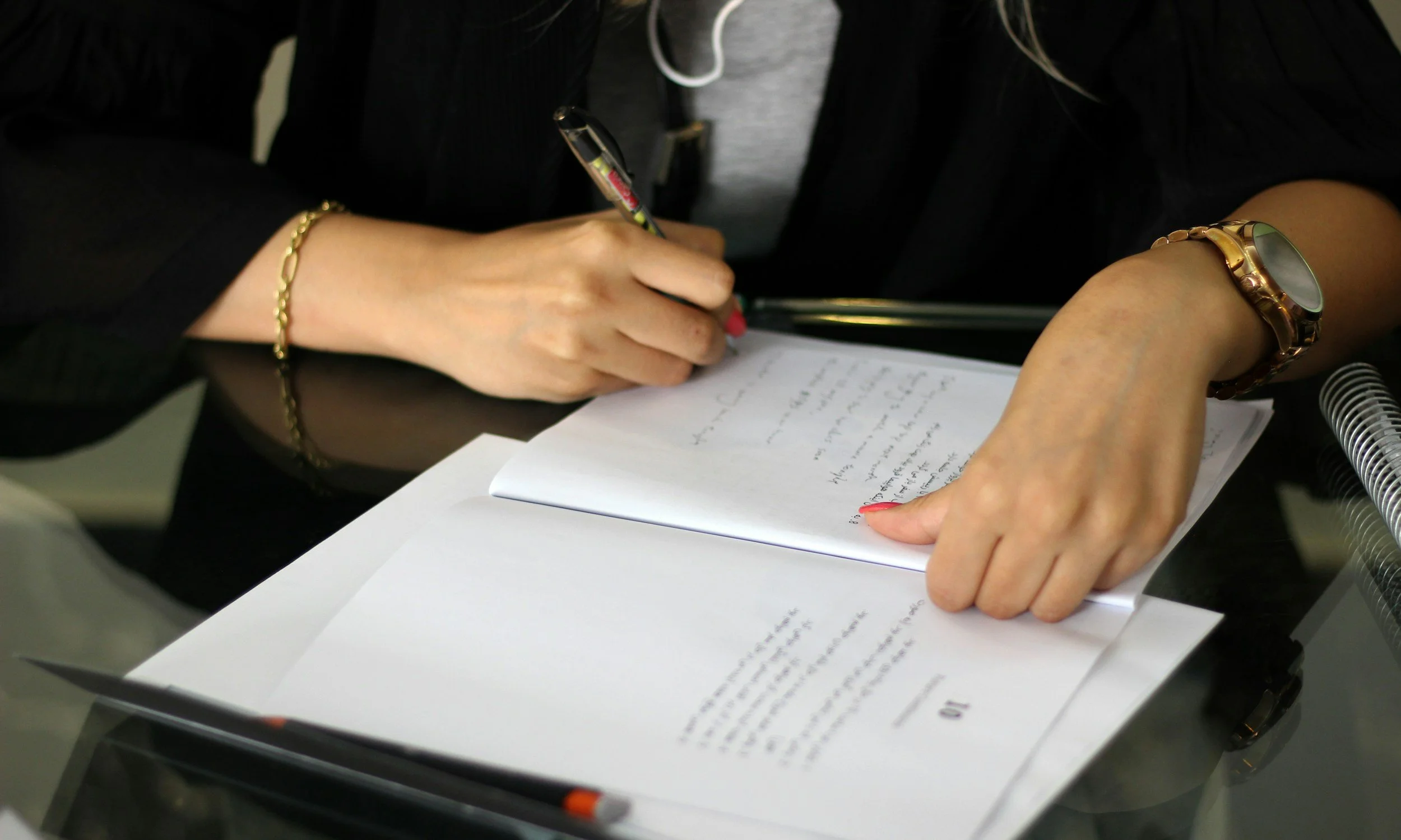 A woman writing on a notepad with a pen, surrounded by papers and documents on a glass table, wearing a watch and a bracelet.