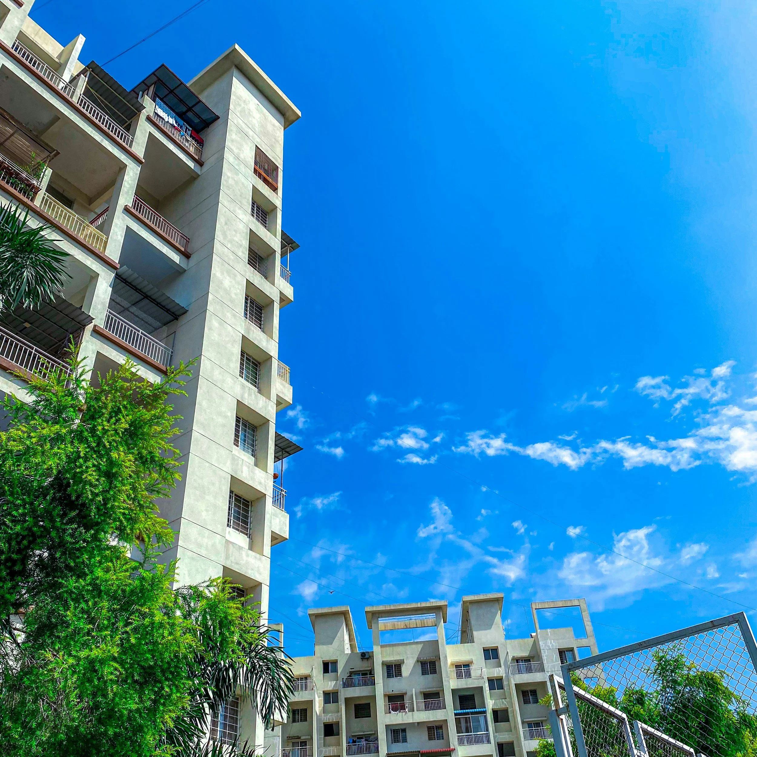 A tall white apartment building with multiple balconies and windows, a blue sky with some clouds, green trees and a metal gate in the foreground.