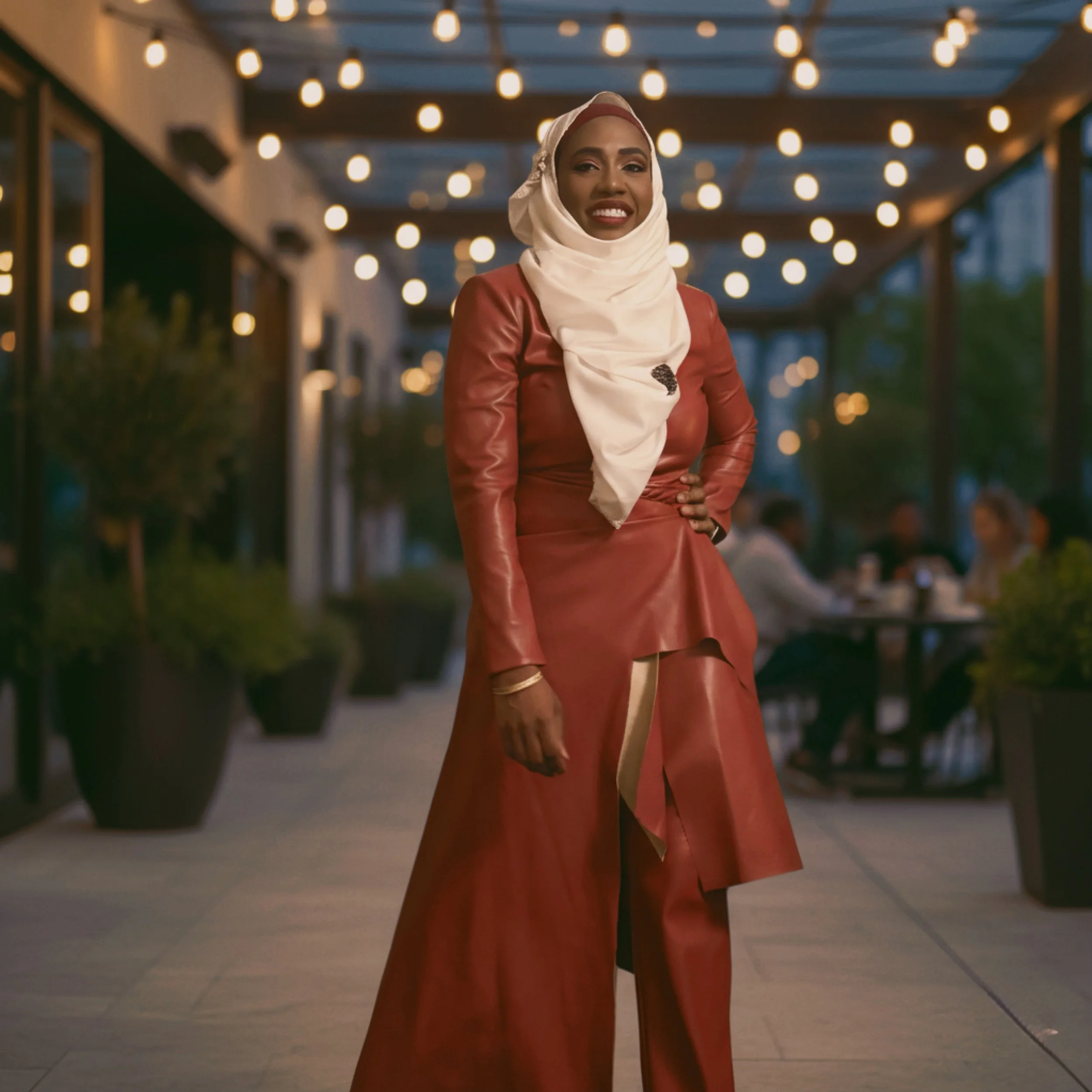 A woman in a red leather dress and a cream-colored hijab posing outdoors at an evening event with string lights overhead.