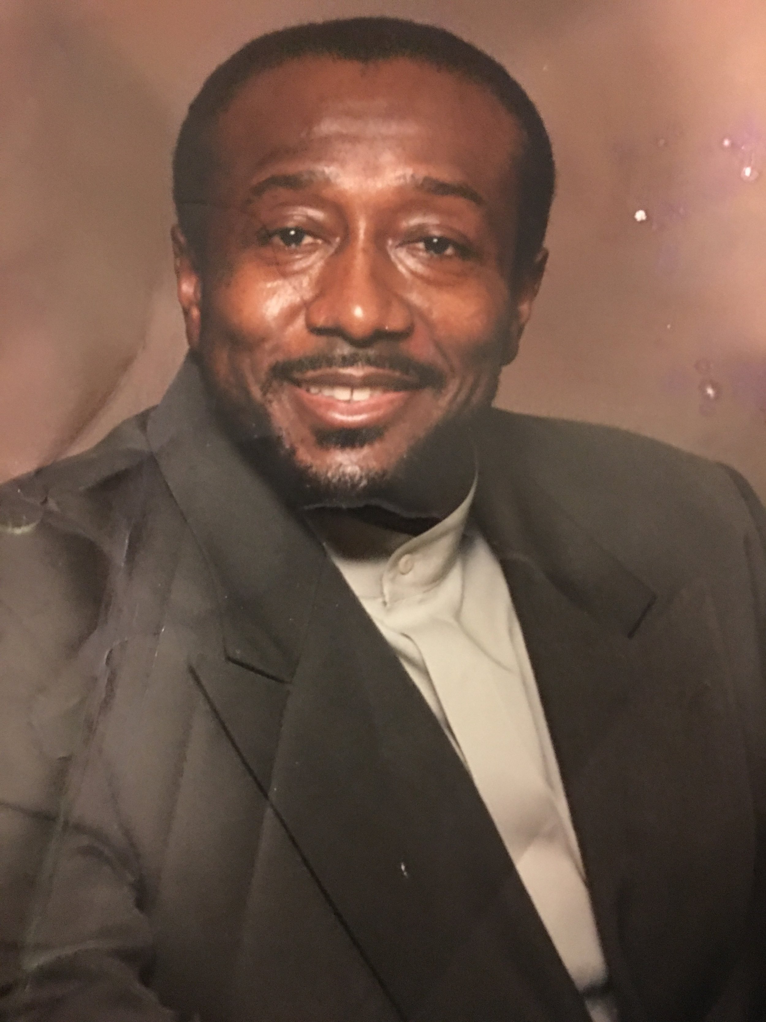 A man with short hair, a goatee, and a mustache, smiling while dressed in a dark jacket and a light-colored shirt, posing for a portrait.