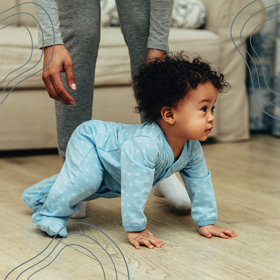 An infant crawling on the floor in blue pajamas with a patterned print, with an adult standing behind, visible from waist down, in a living room with a beige couch in the background.