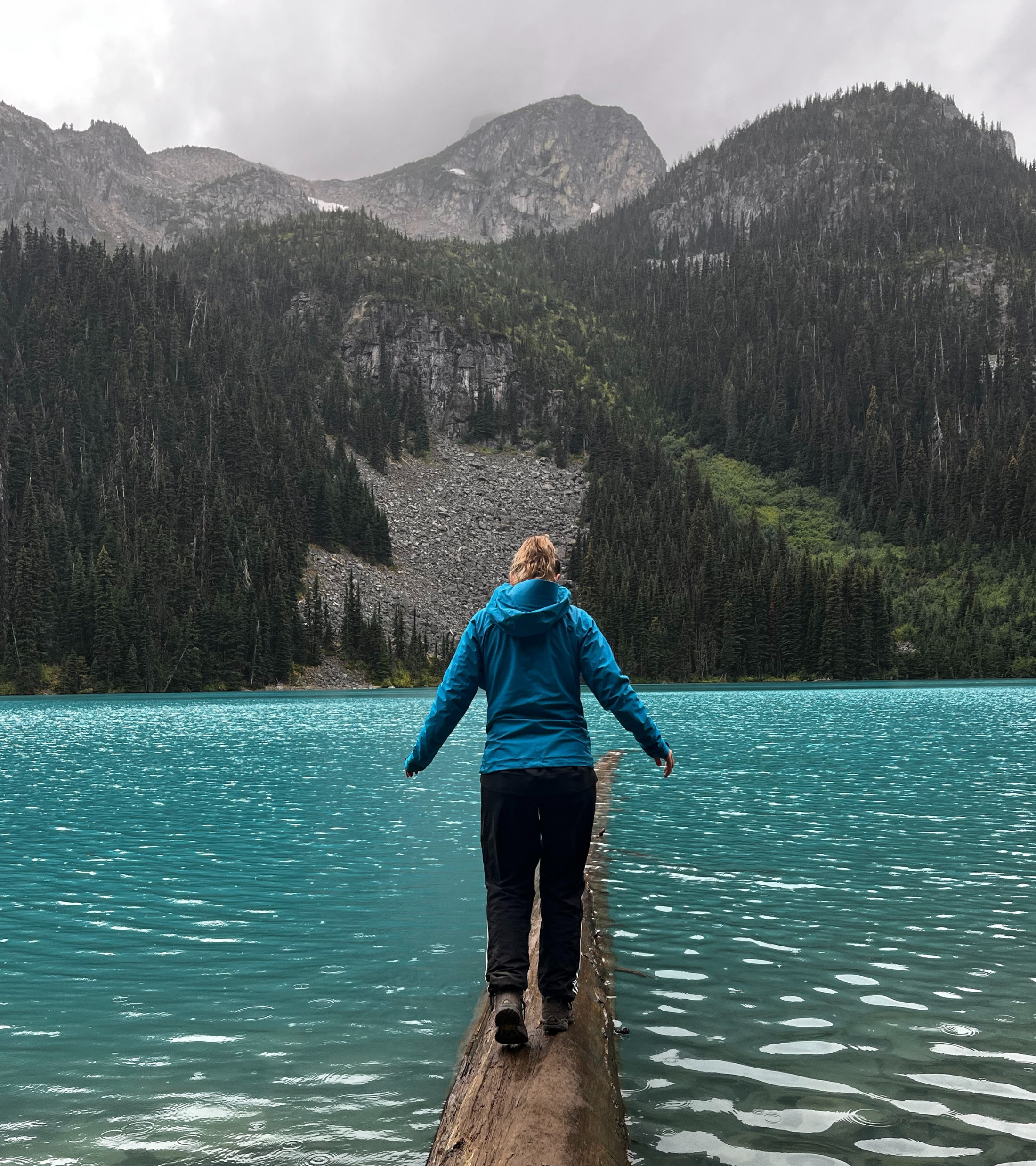 Person in blue jacket walking on a log in a lake with forested mountains and cloudy sky in the background.