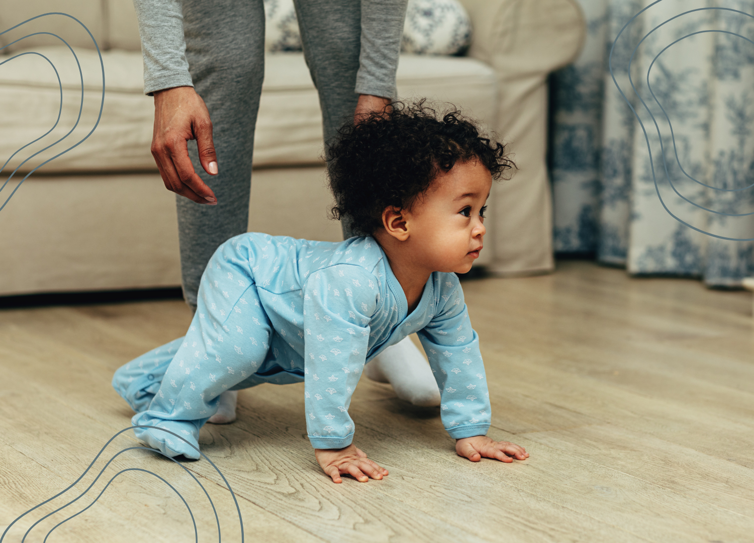 An infant crawling on the floor in blue pajamas with a patterned print, with an adult standing behind, visible from waist down, in a living room with a beige couch in the background.
