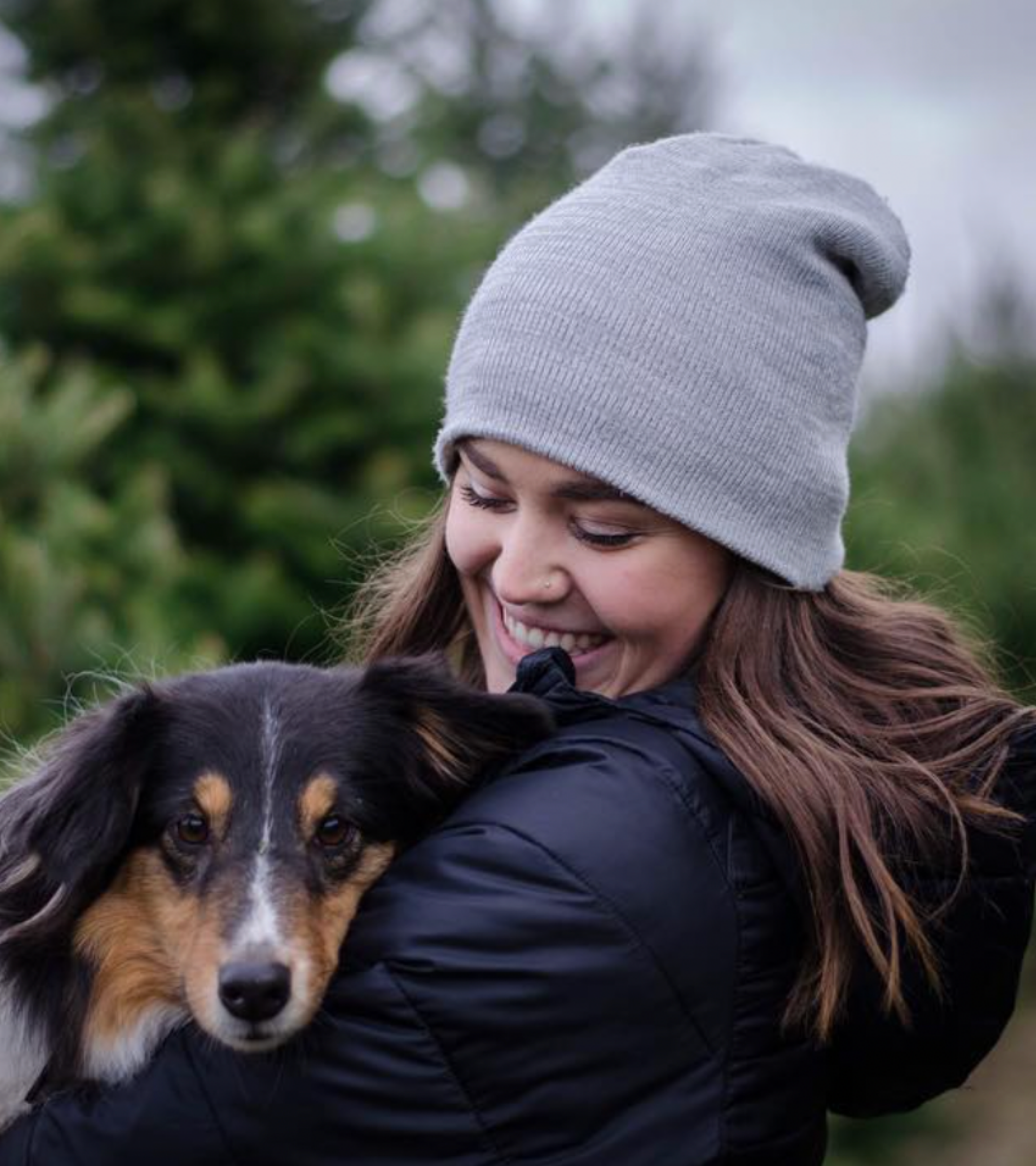 A smiling young woman with brown hair wearing a gray beanie hat, holding a black and brown dog outdoors.