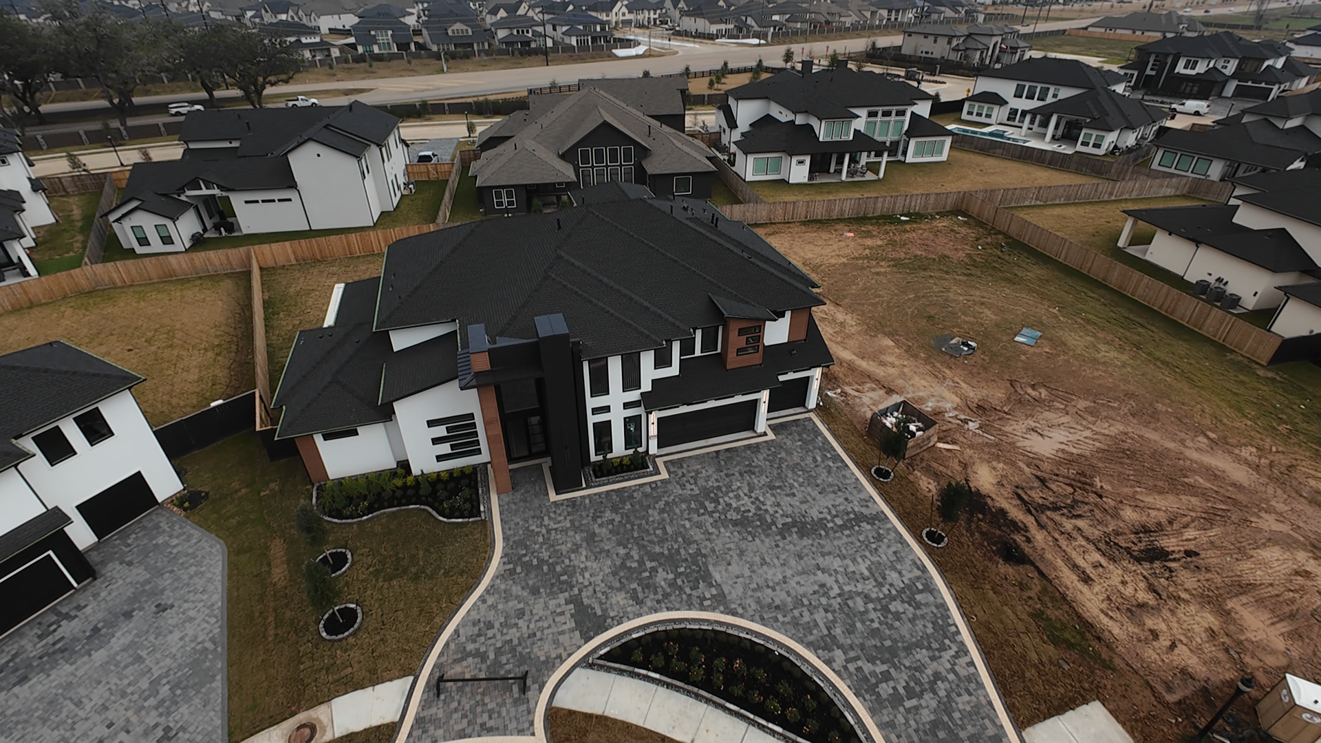 Aerial view of a newly built modern house with a black roof, white and brown exterior walls, a black garage door, and a paved driveway. Surrounding plots of land and neighboring houses in a suburban neighborhood.